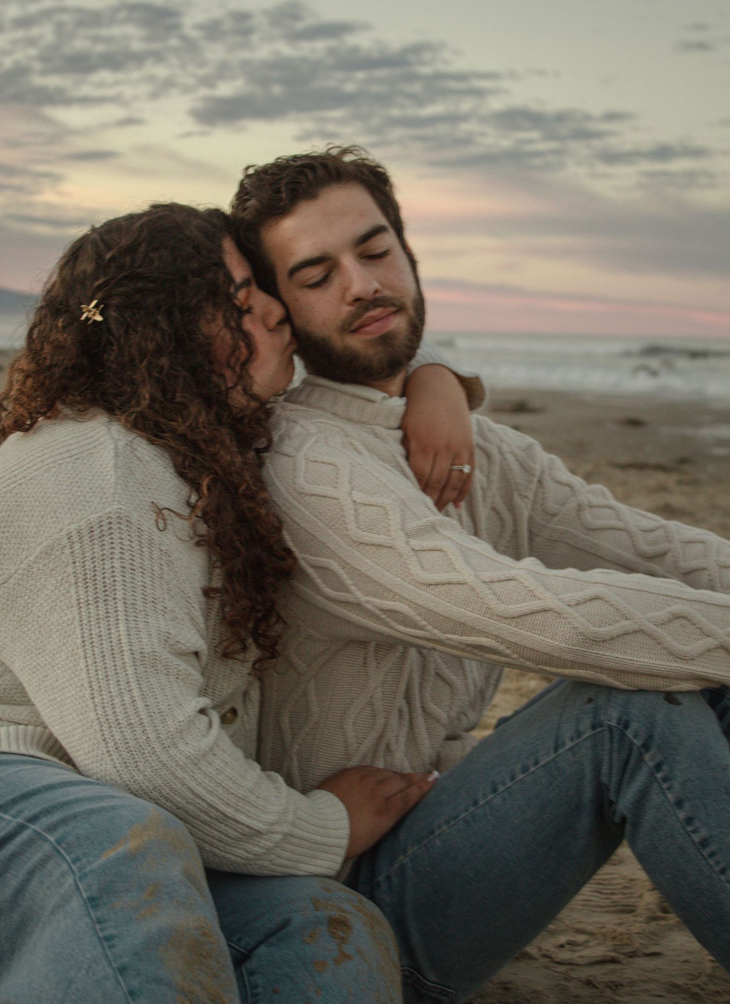 A couple in matching cream cable knit sweaters share an intimate moment on the beach at sunset.