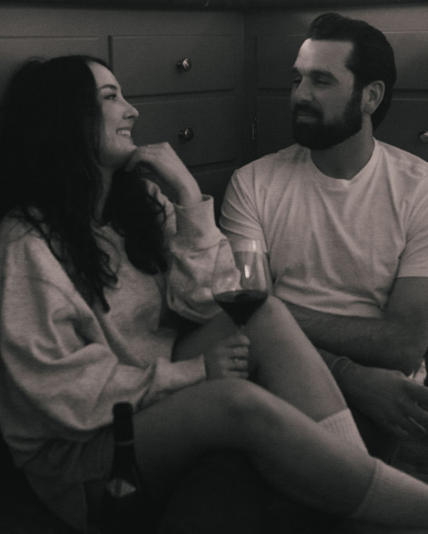 Black and white photo of a smiling couple sitting together, woman laughing while man holds a glass of red wine.