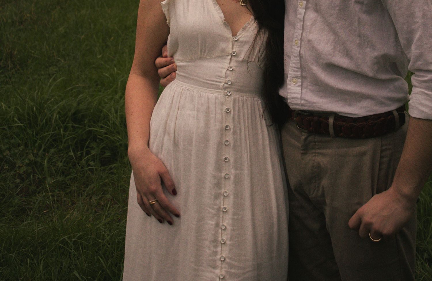 A couple stands together on grass, with focus on vintage-style white dress details and button details.