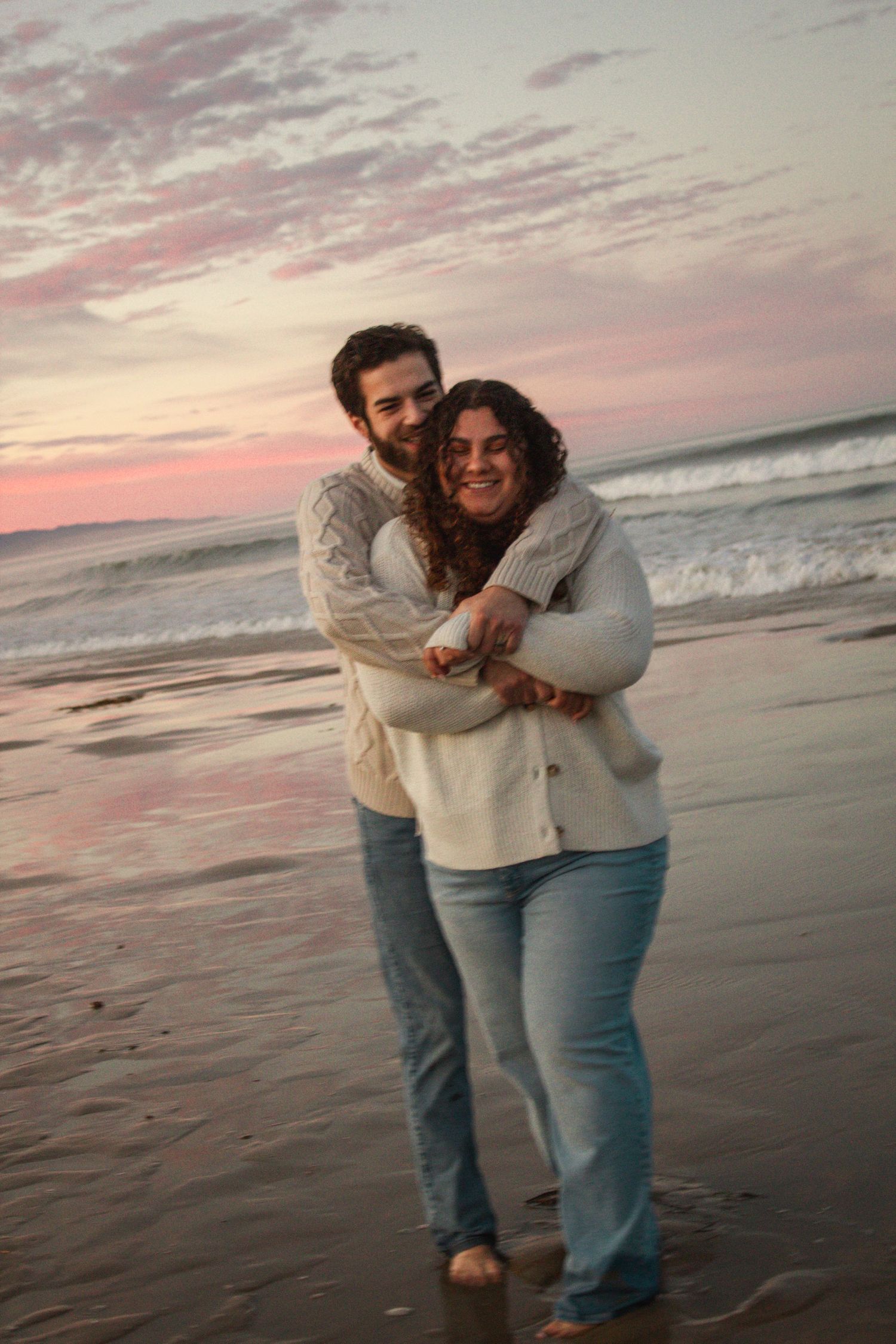 A couple shares a tender moment on the beach at sunset with pink and purple skies reflecting on the ocean waves.