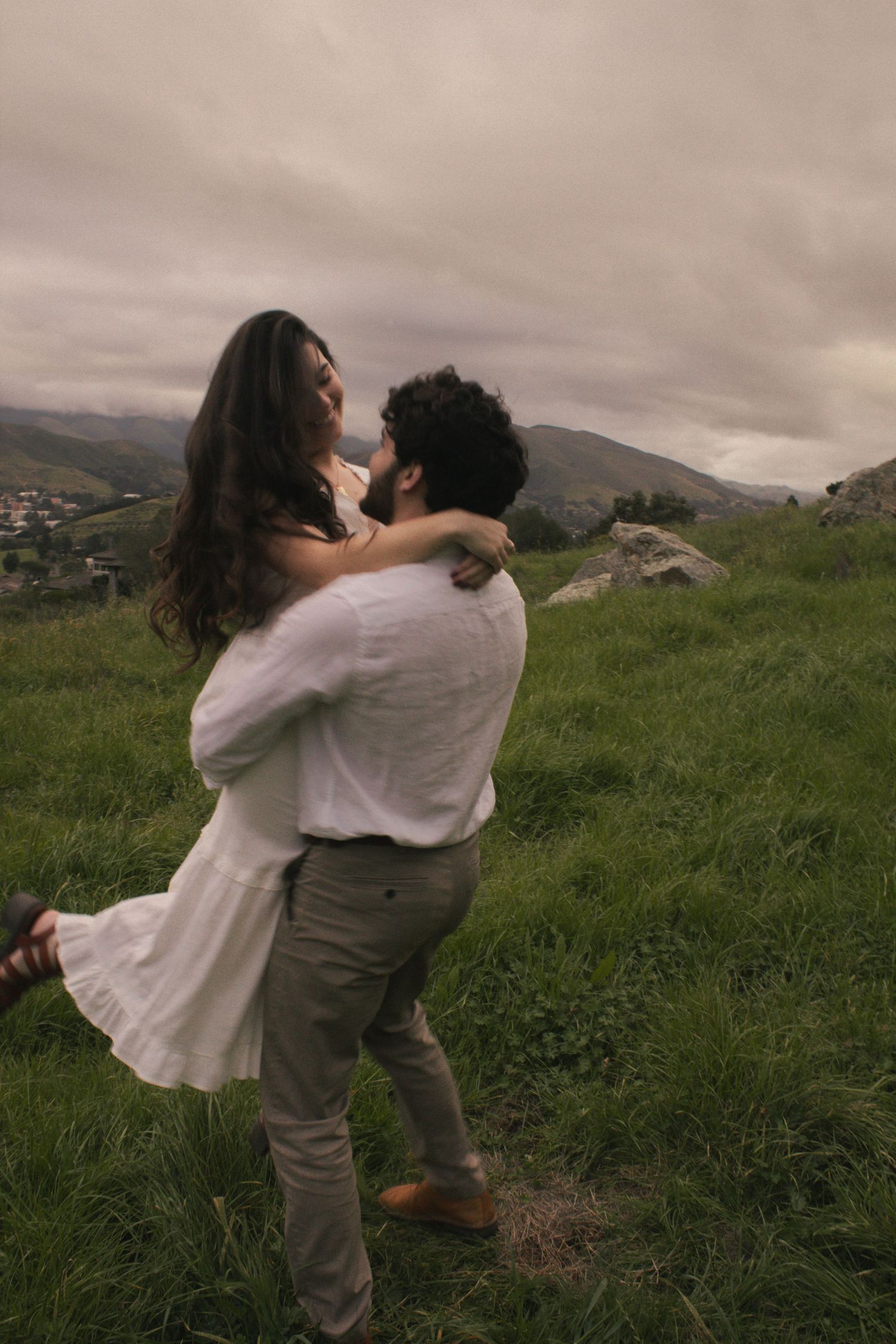 A couple in white and gray clothing embrace while standing in a lush green hillside meadow under moody skies.