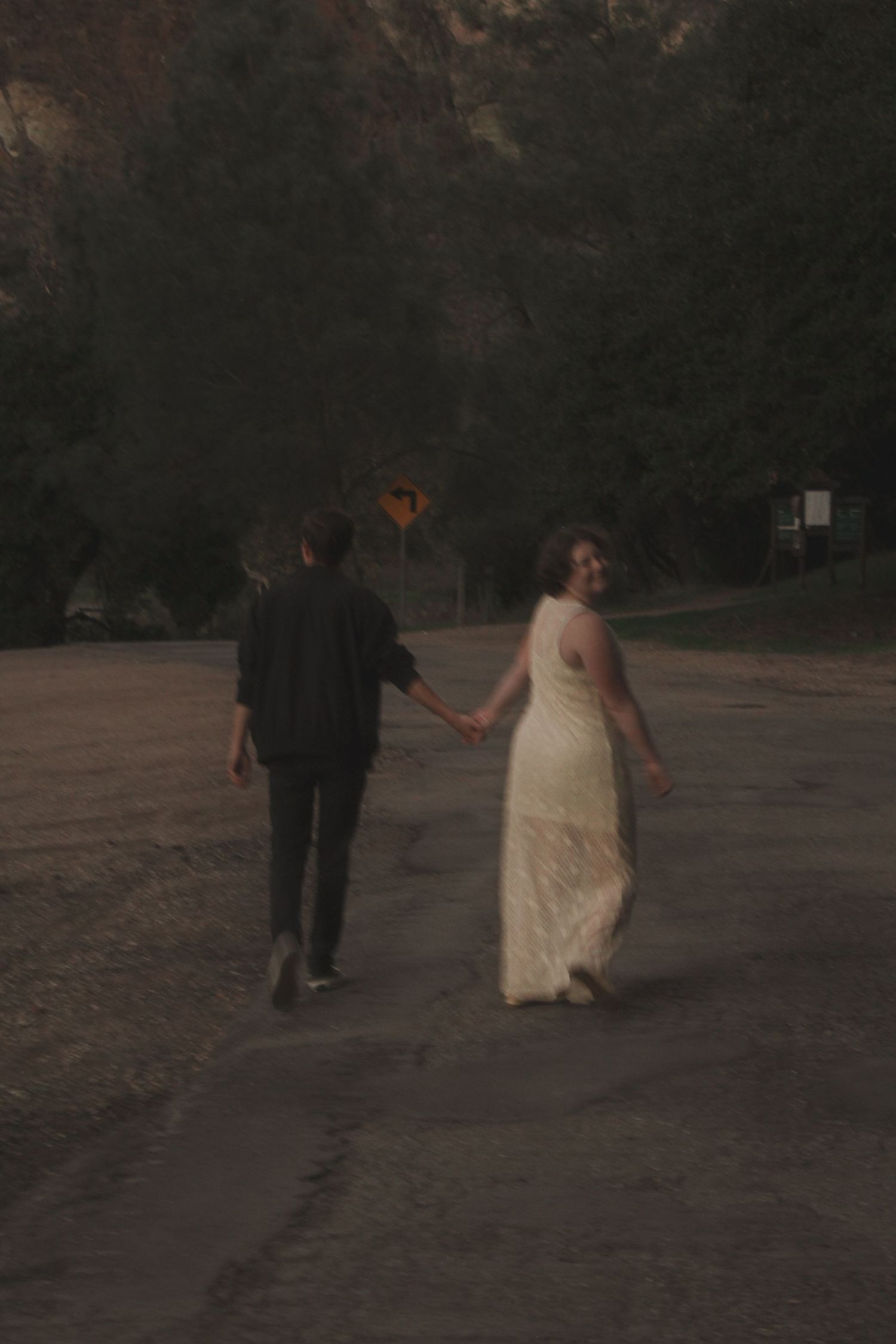 A couple in wedding attire walks away from the camera along a dark path in a park setting.