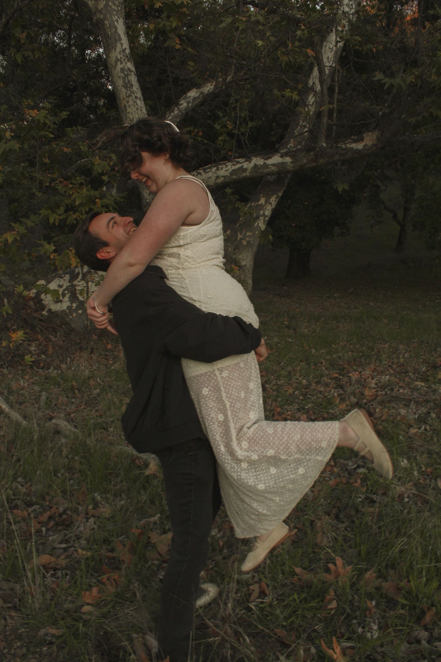 A romantic outdoor embrace shows a couple in formal wear sharing a playful lift pose against a wooded backdrop.
