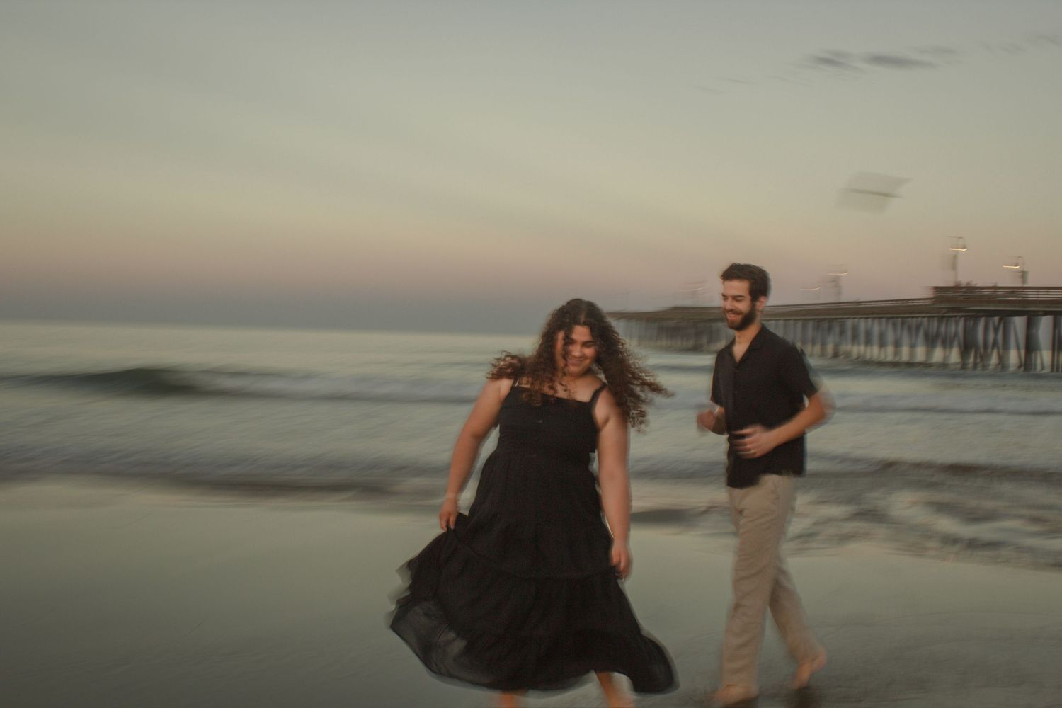 A couple shares a romantic moment on a misty beach at sunset with a pier in the background.