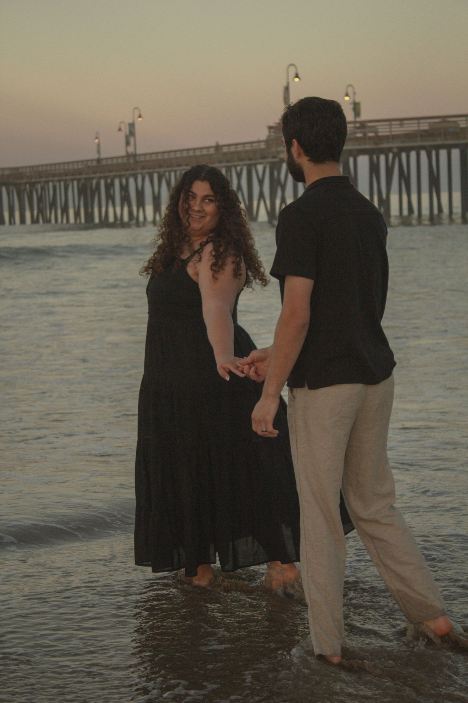 Couple wading in shallow ocean water at sunset near a pier, creating romantic silhouettes against the golden light.