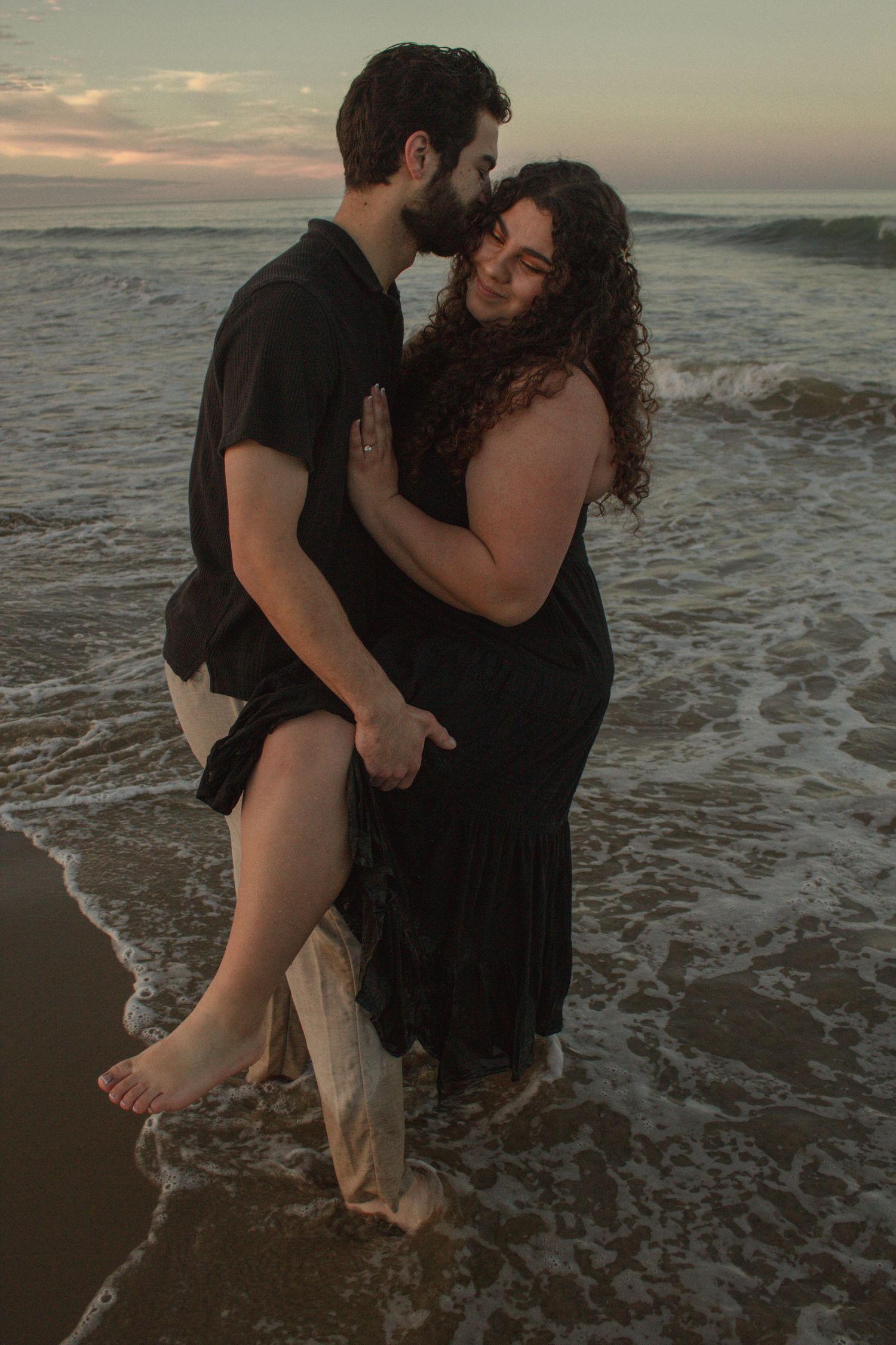 A couple shares a romantic moment on the beach at sunset as waves crash around their feet.