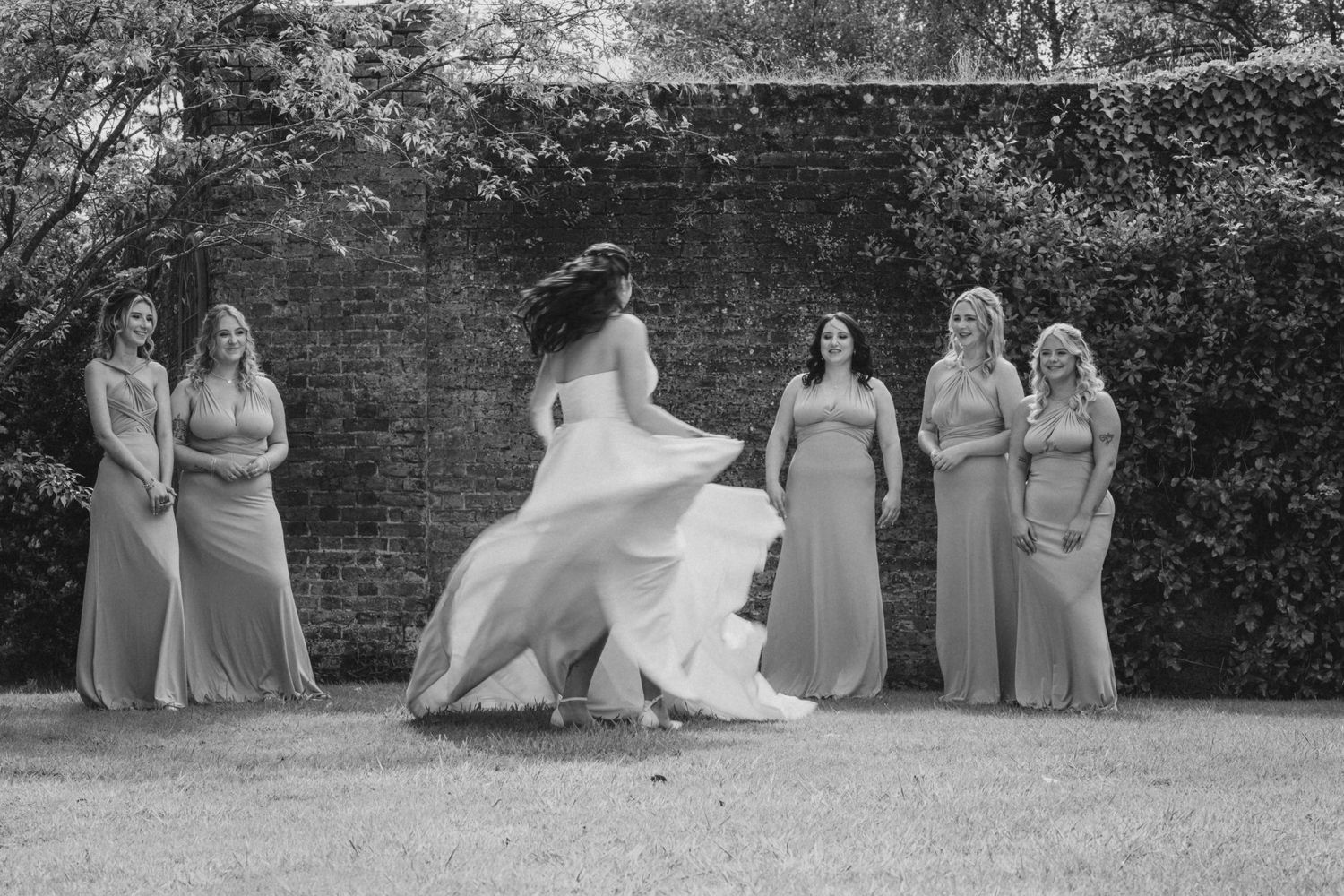 Wedding party posing on grass in front of brick wall backdrop.