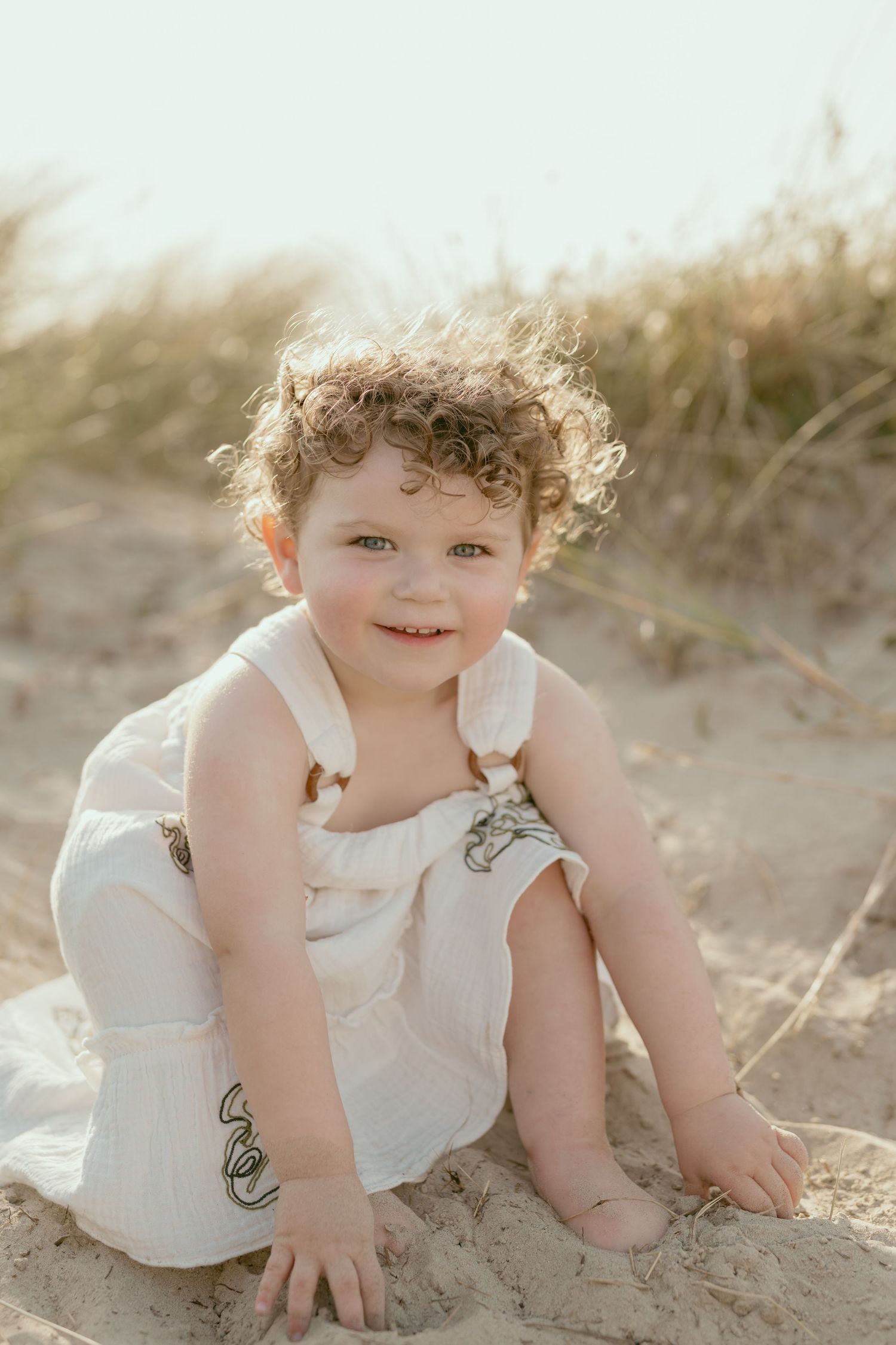A small child wearing a white sundress crawls on sandy beach during sunset.
