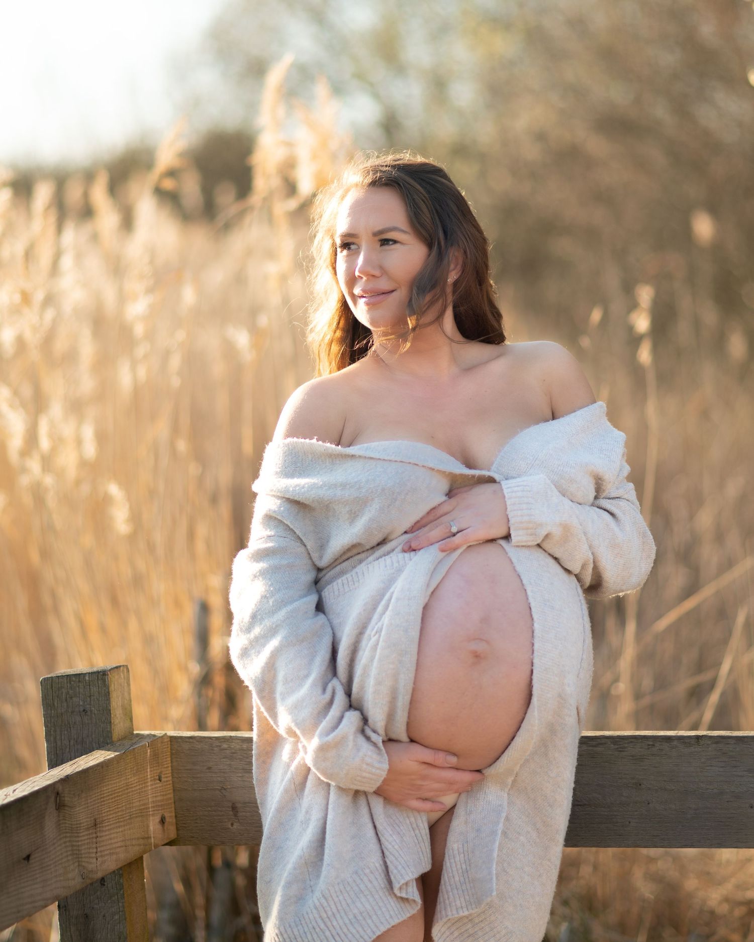 Maternity photos in golden light showing pregnant figure in cozy white sweater on wooden fence.