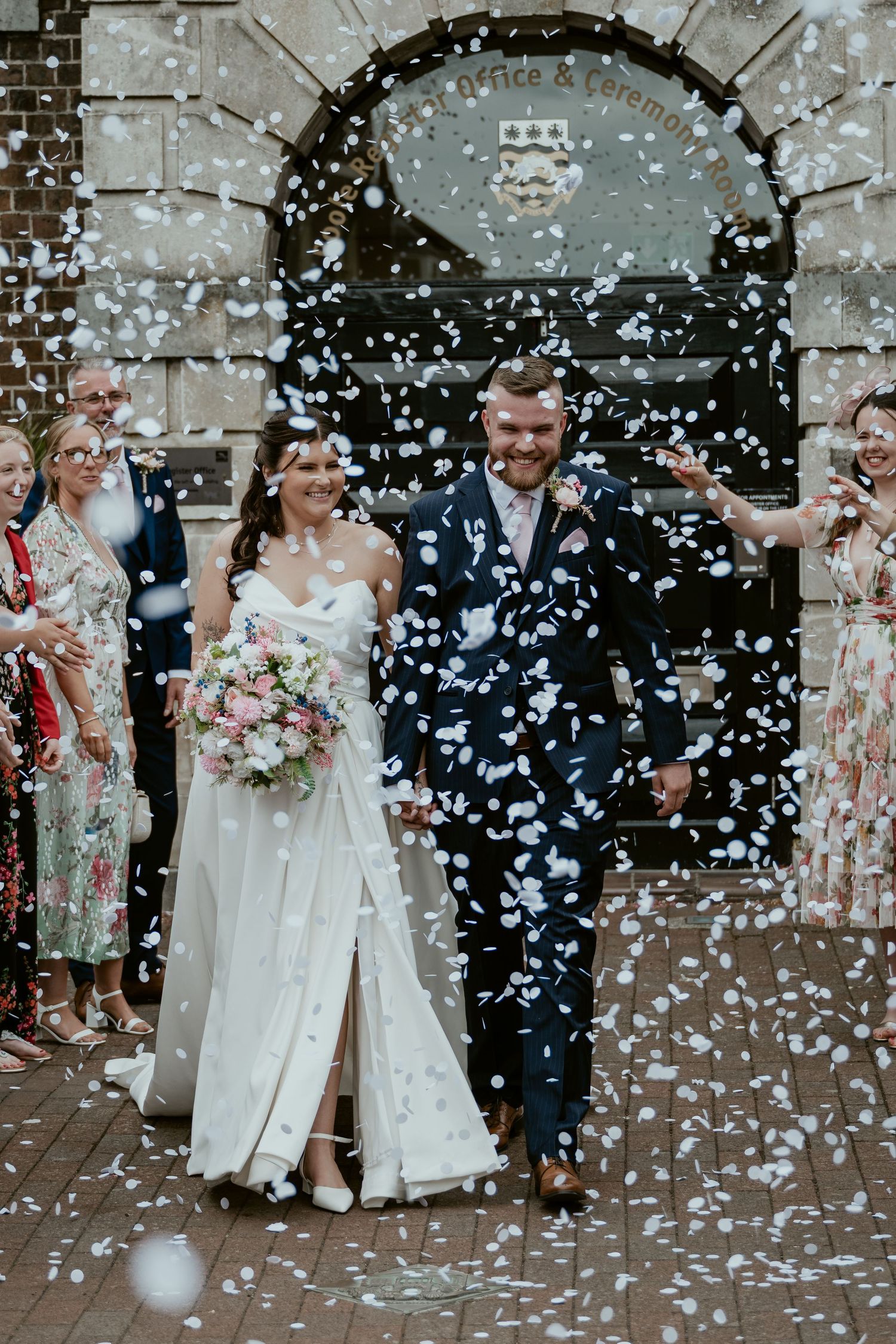 Wedding party celebrates under a shower of white confetti outside a historic building.