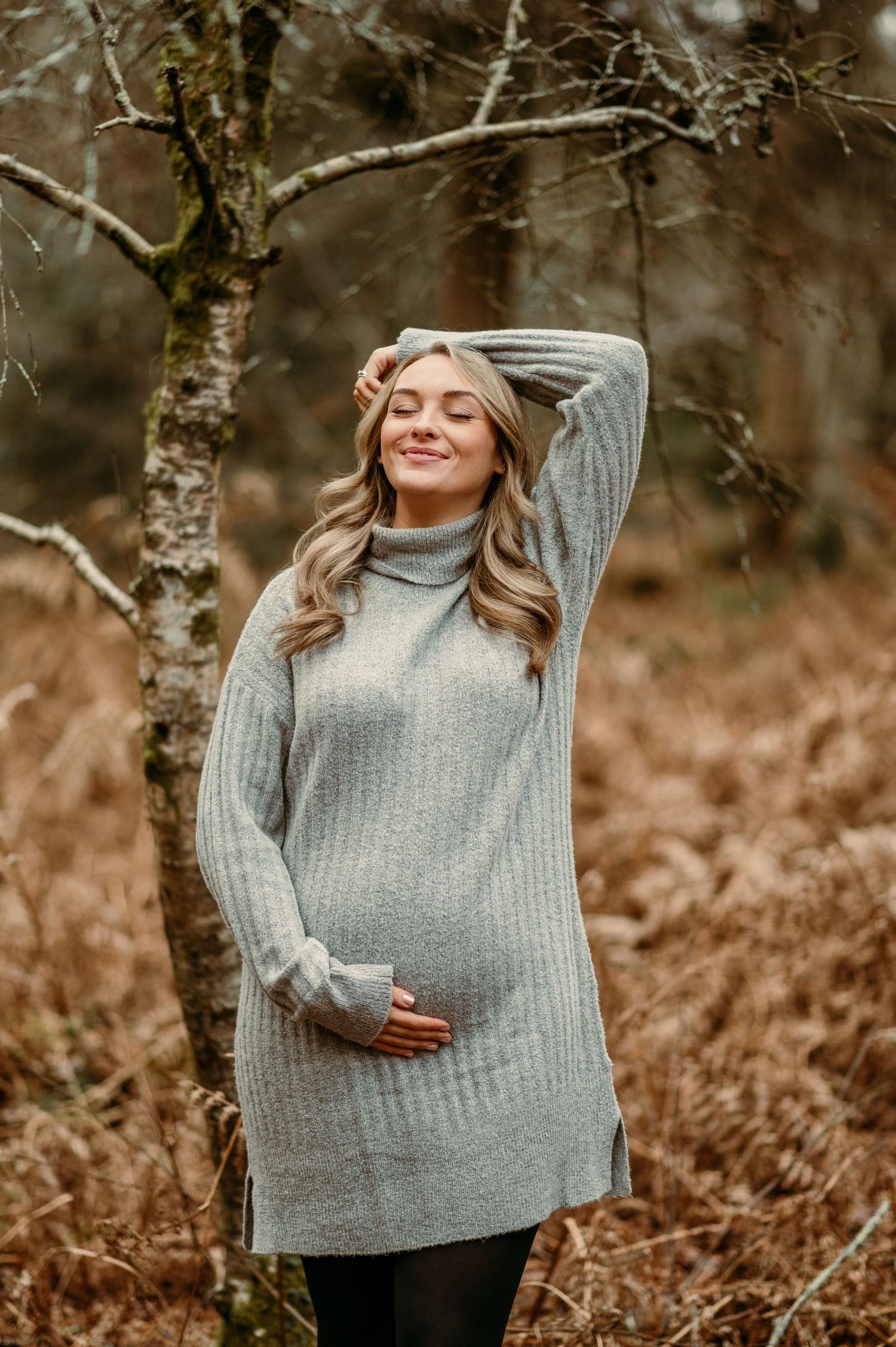 Maternity portrait of someone wearing a grey sweater against autumn foliage.