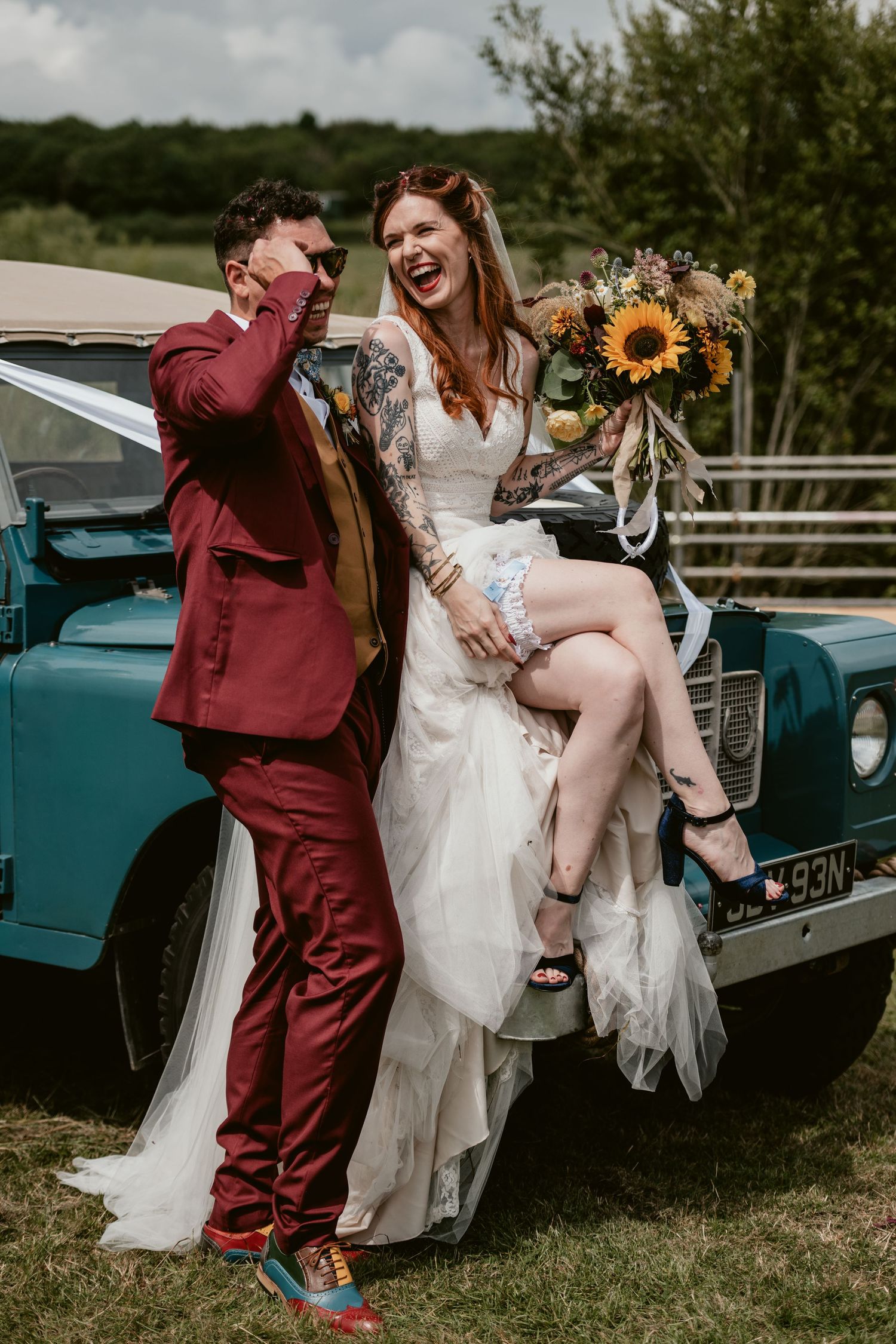 Wedding party poses playfully by vintage turquoise car with sunflowers in background.