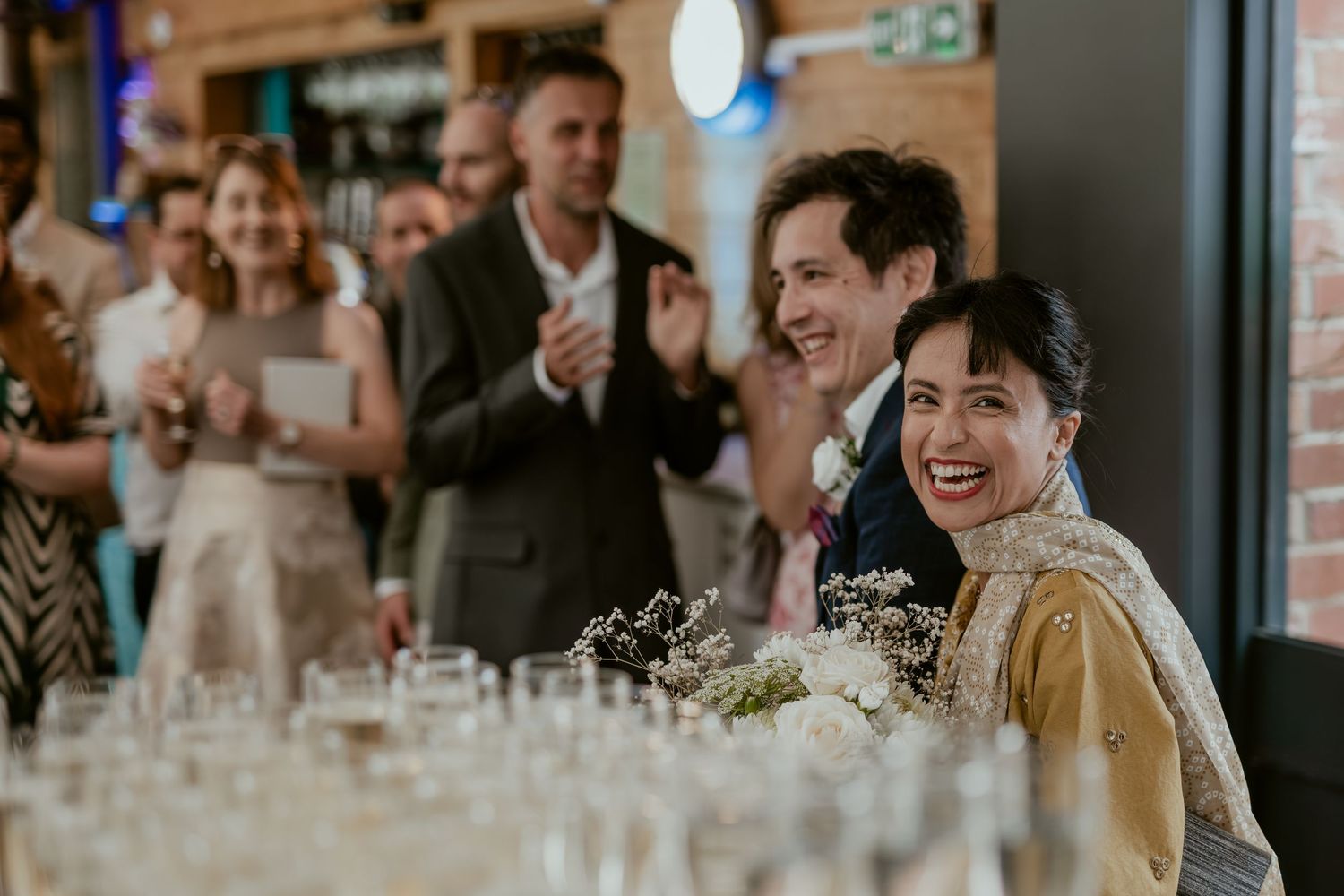 Wedding reception scene with guests celebrating at wooden tables in rustic venue.