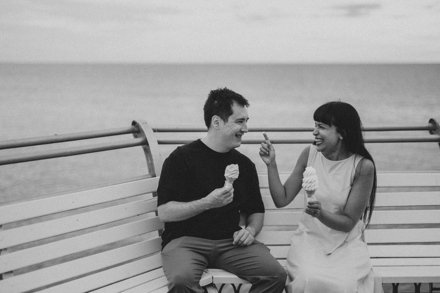 A couple enjoys ice cream together on a waterfront bench during an overcast day.