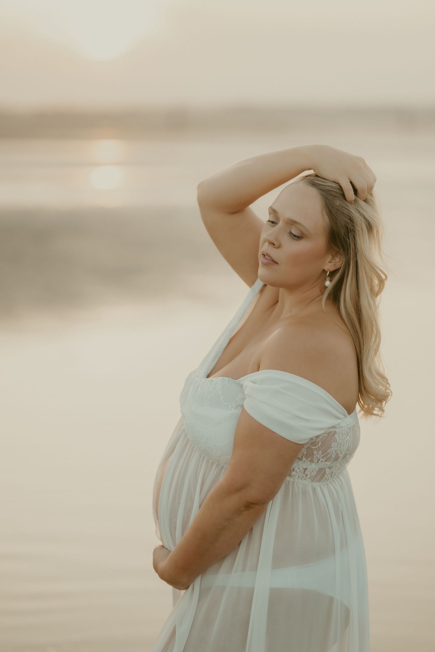 Ethereal maternity photo of woman in white dress at sunset by water.