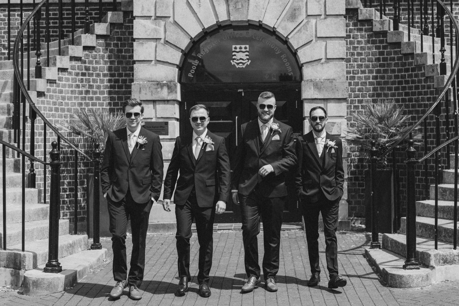 Wedding party in formal attire poses outside a brick building entrance.