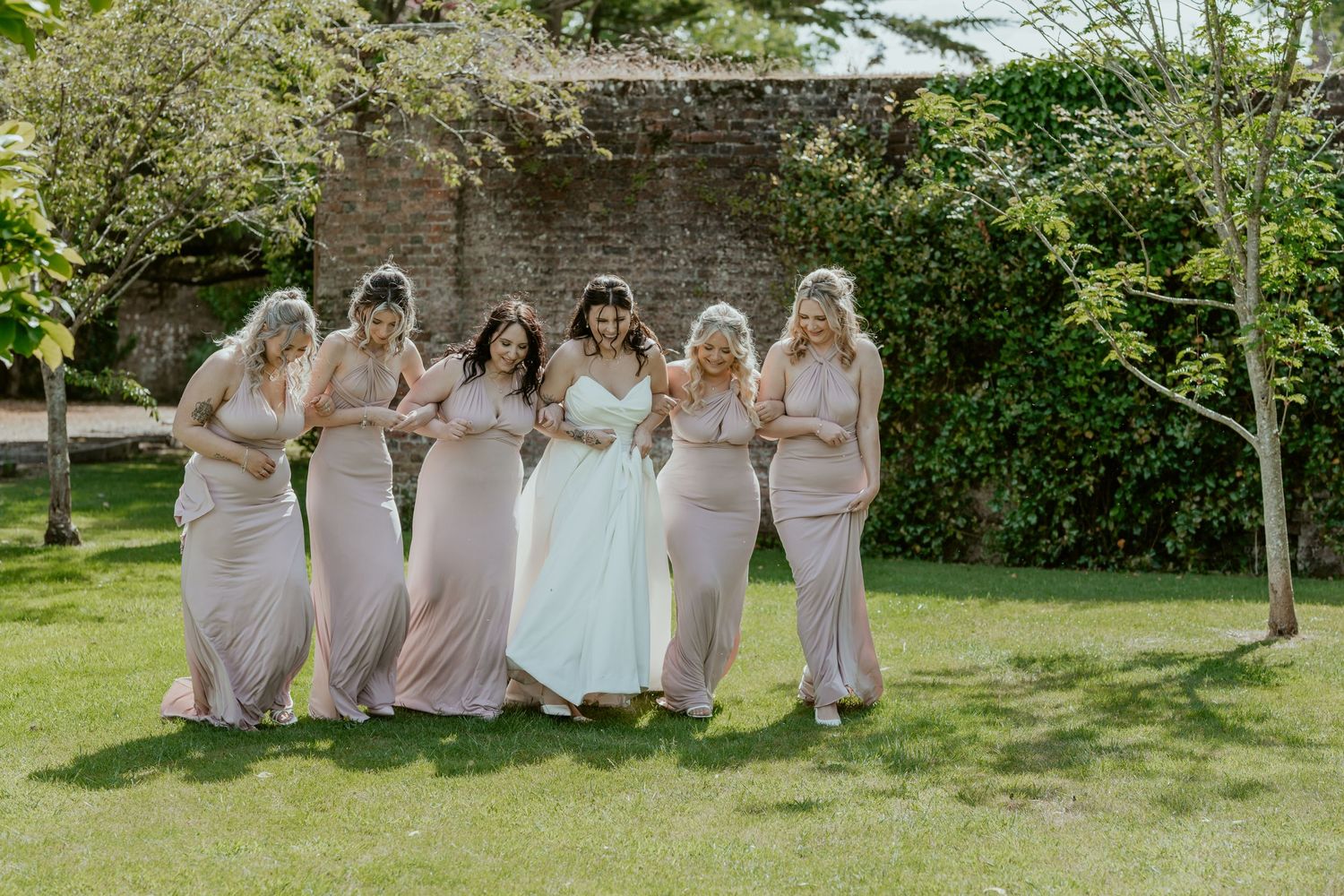 Bridesmaids in blush pink dresses pose together on a sunny lawn.