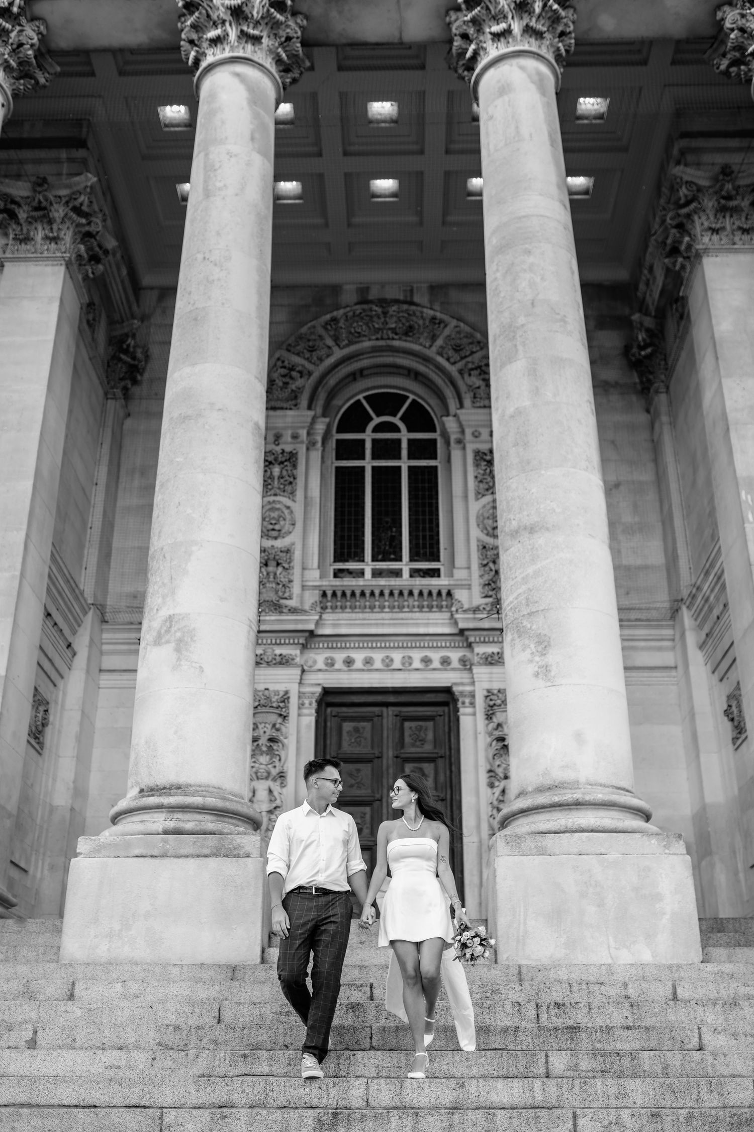 Couple in white clothing standing between classical columns of historic building.