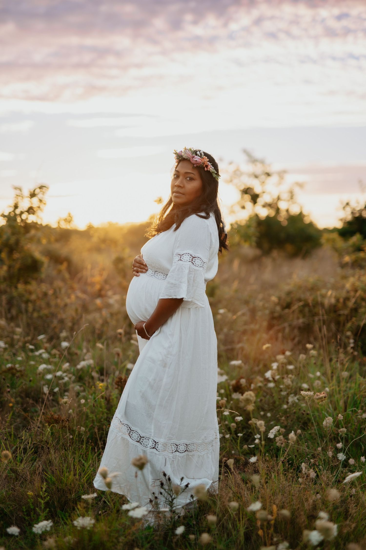 Maternity portraits in flowing white dress amid field of wildflowers during golden hour.