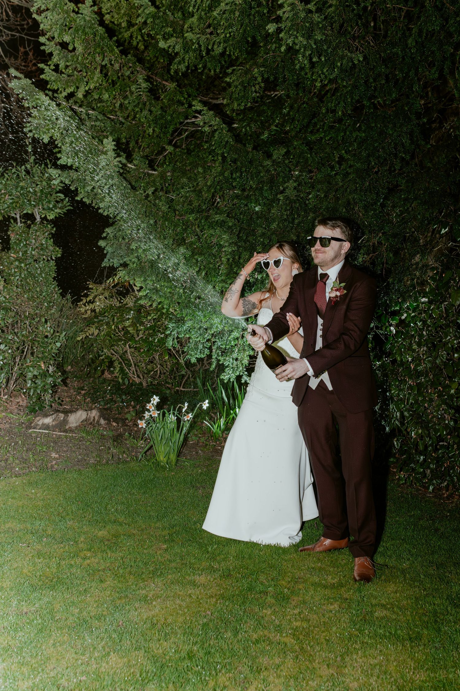 A couple in formal wedding attire dances together in a garden setting at dusk under green foliage.