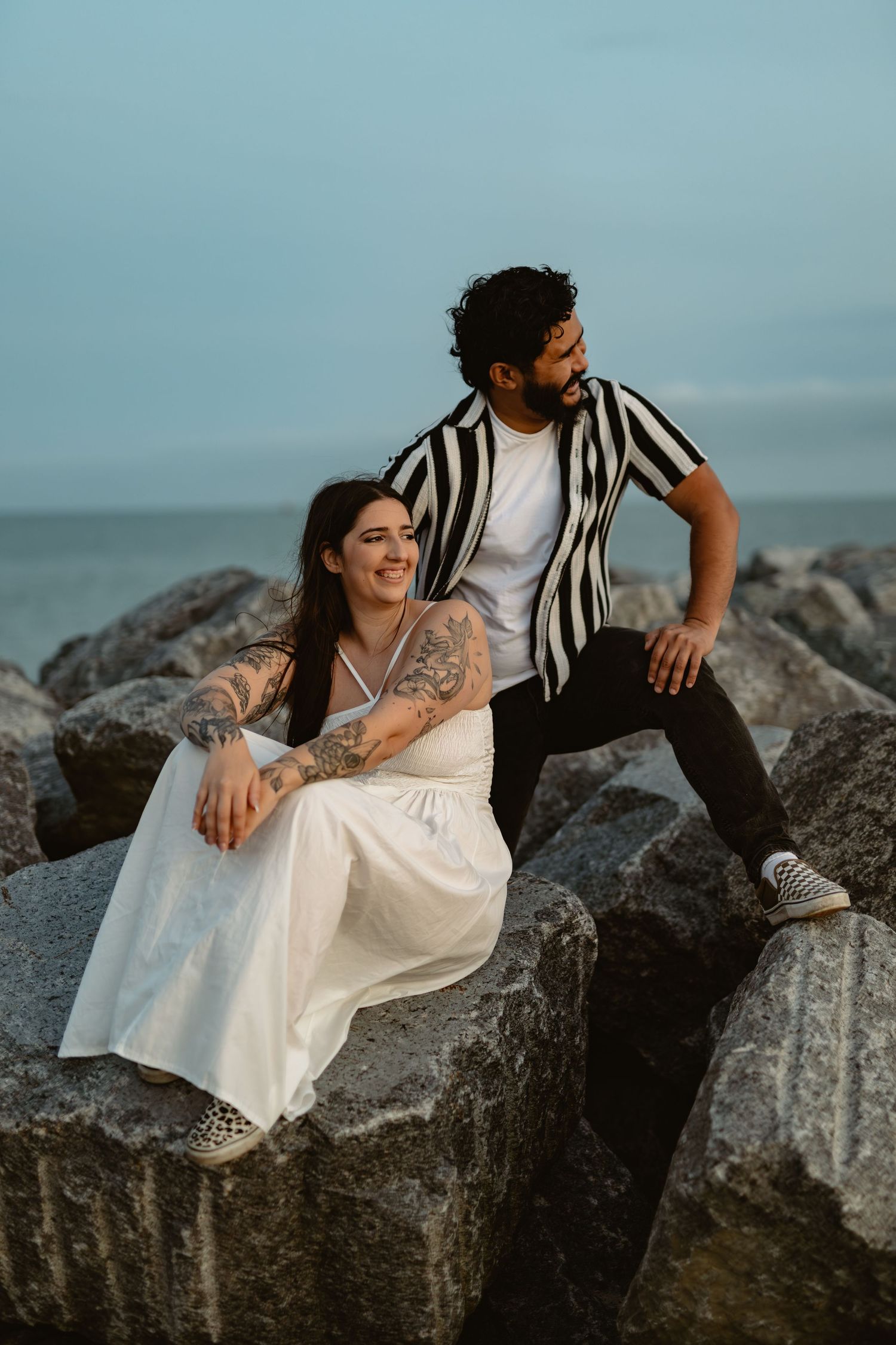 Two people relax on rocky coastal formations against a moody blue sky backdrop.