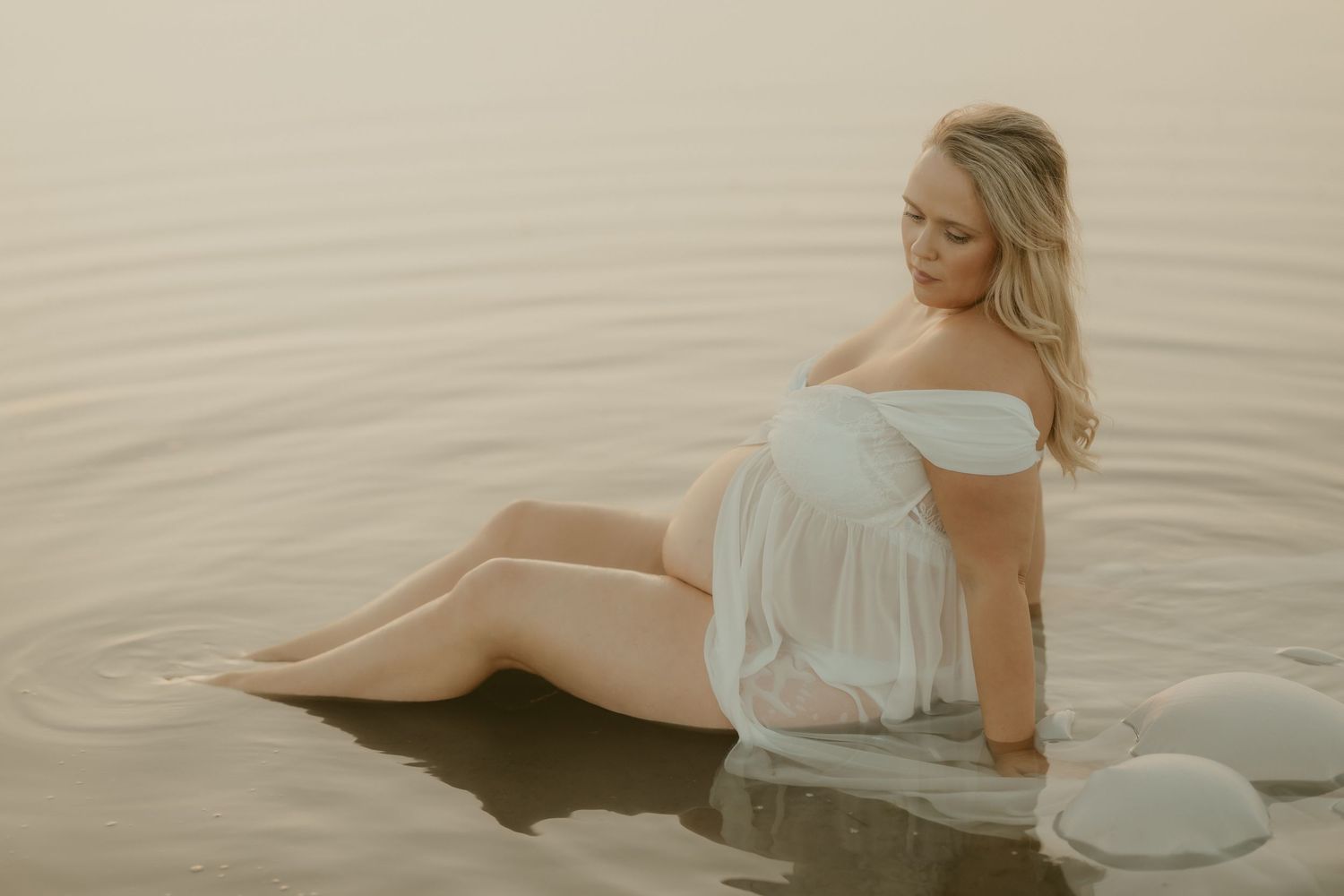 Woman in white dress lying gracefully in shallow beach water during sunset.