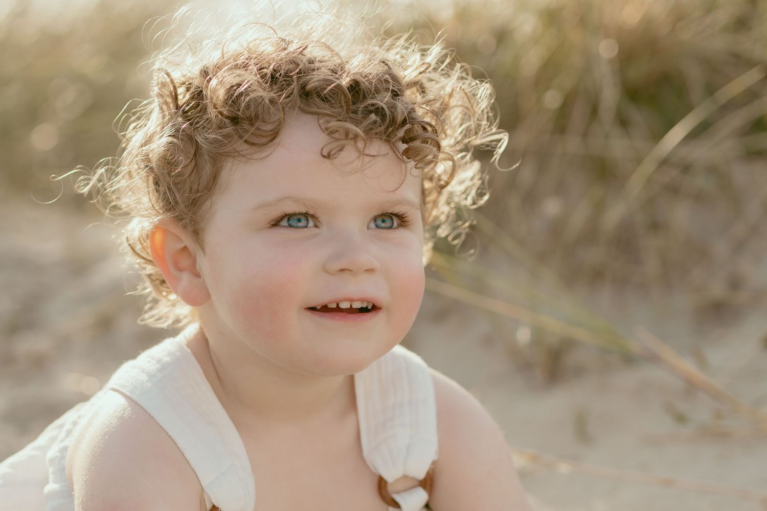 A toddler in a white dress with curly hair plays on the beach in golden sunlight.