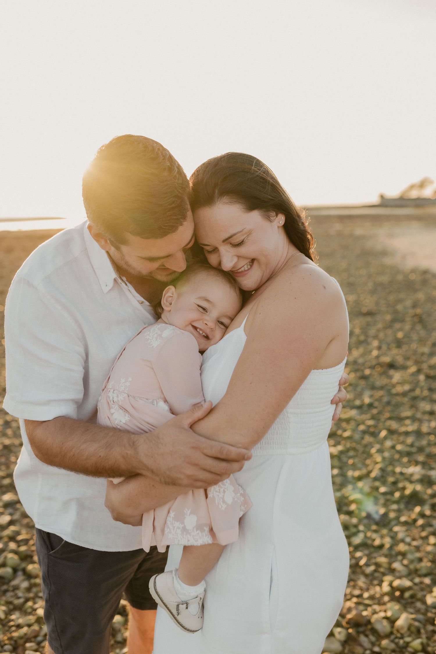 Family sharing tender moment on beach at golden hour in white outfits.