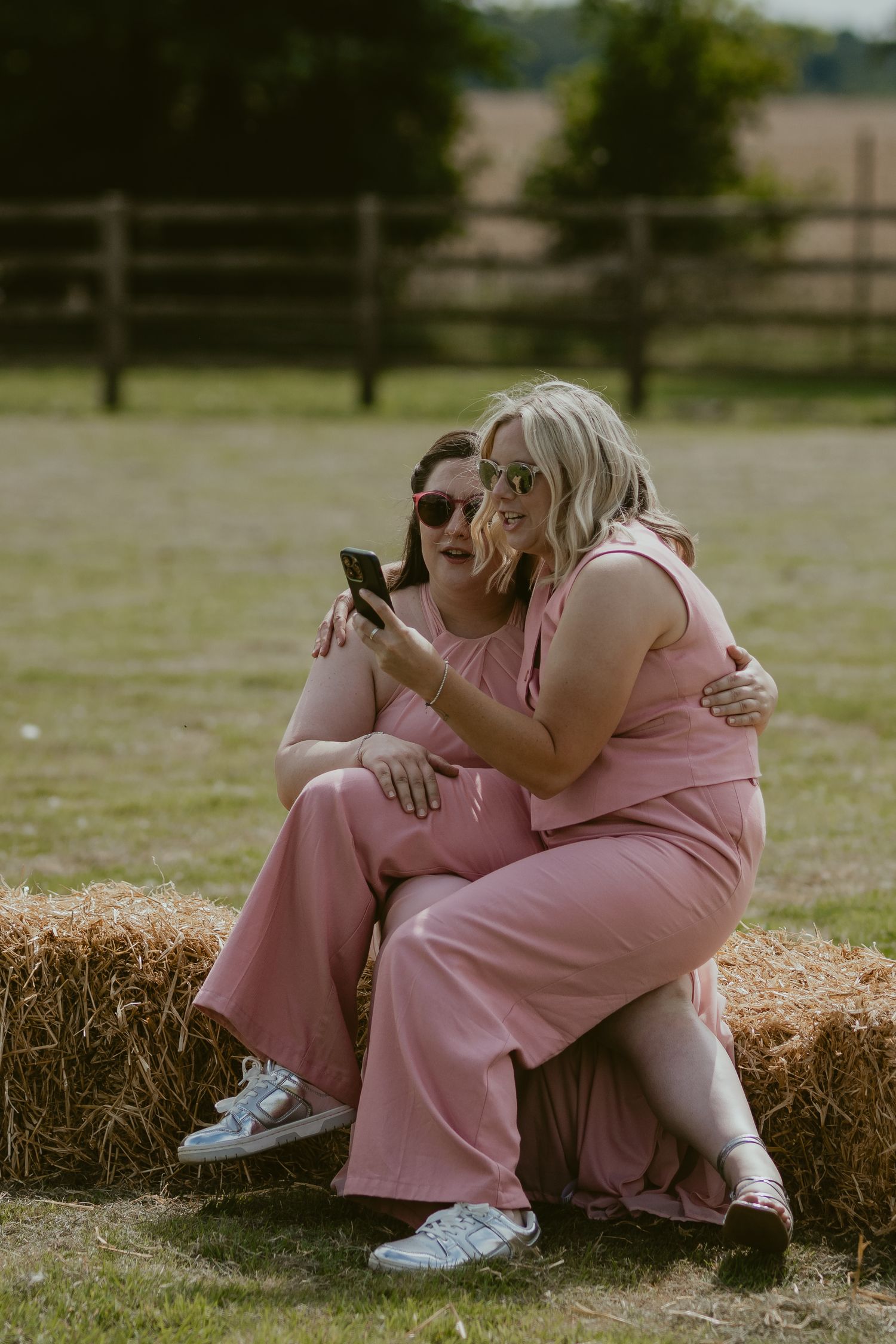 Two guests in pink outfits sitting on hay bale looking at phone.