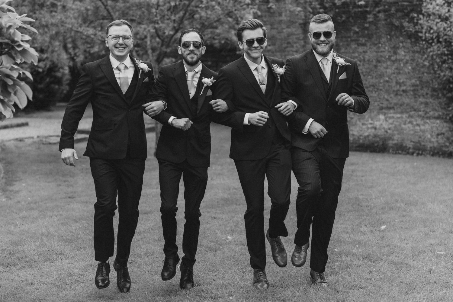 Groomsmen in black suits running playfully across a garden path.