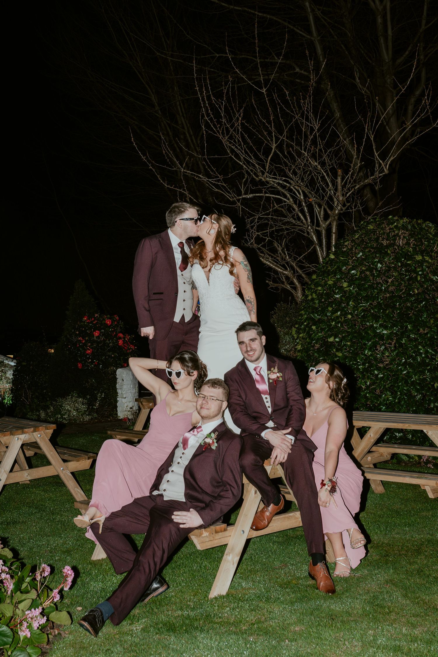 Wedding party members in burgundy suits and pink dresses pose playfully on wooden picnic tables at dusk.
