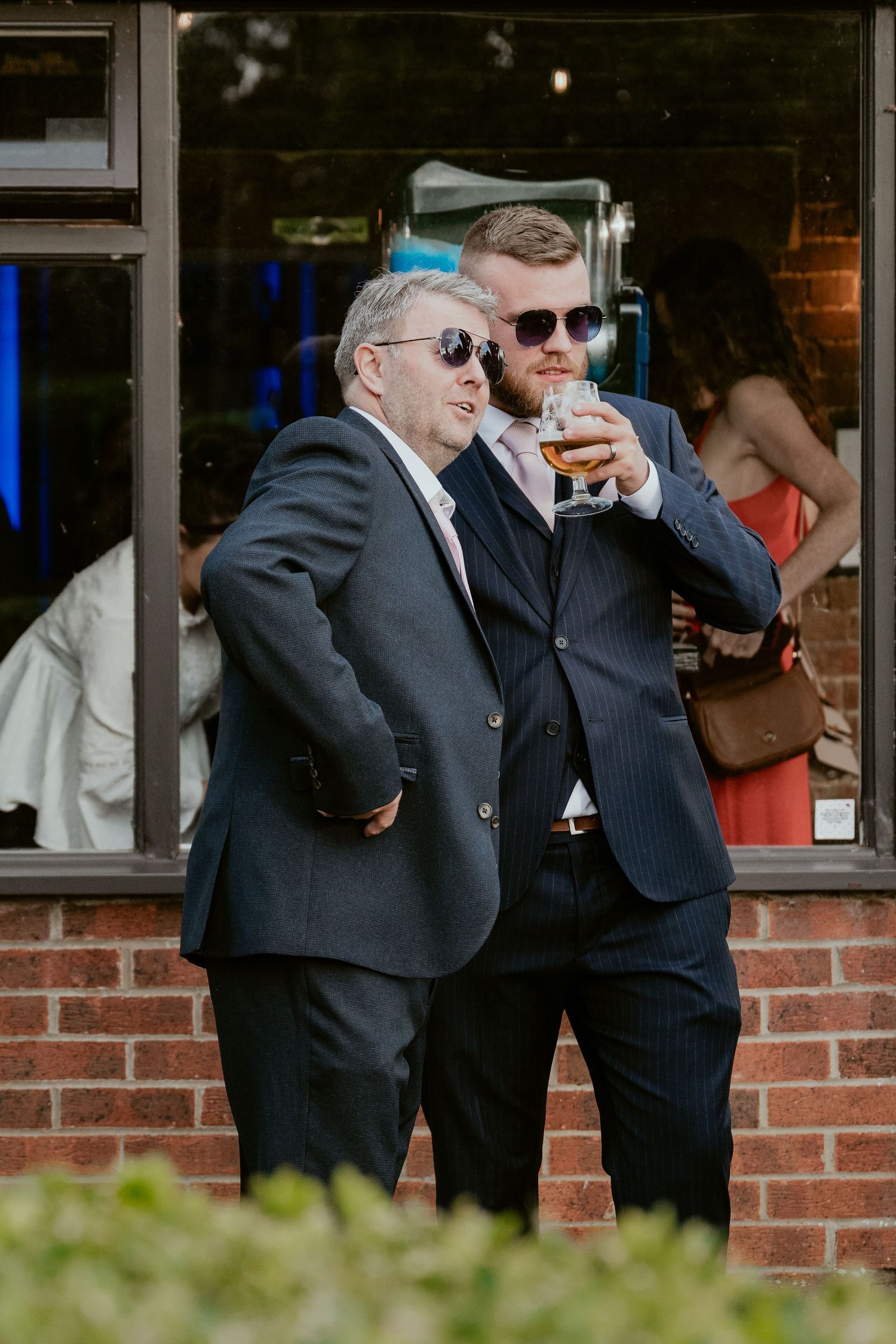 Groomsmen in dark suits share drinks during outdoor wedding festivities.