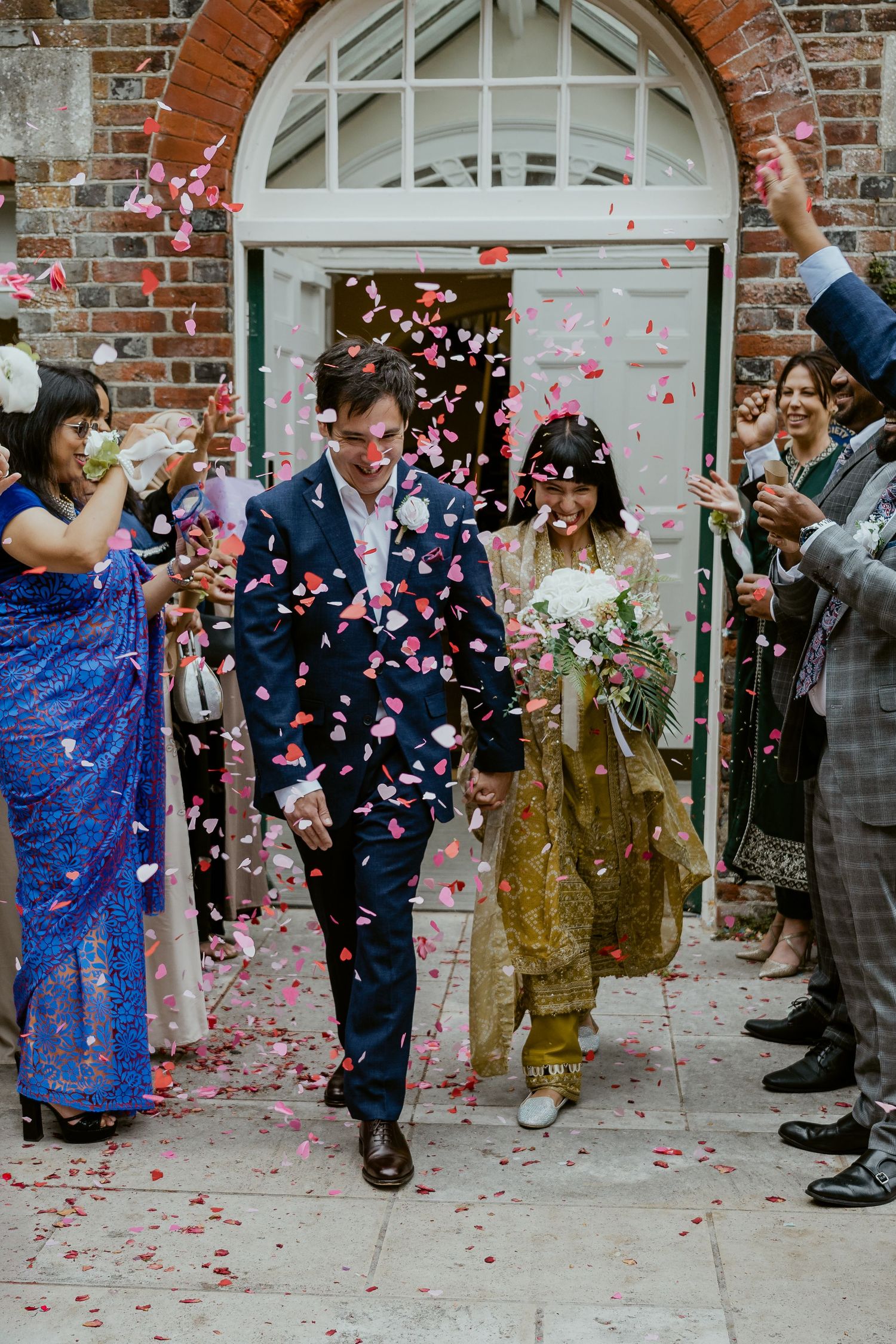 Wedding guests shower a couple with pink confetti as they exit through a brick archway entrance.