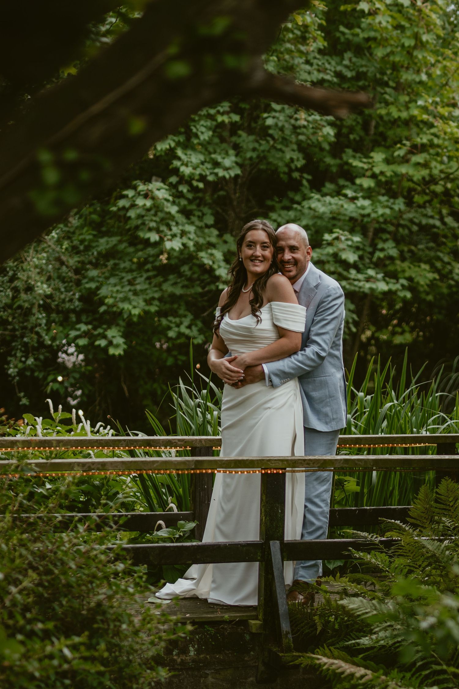 Newlyweds embrace on a wooden bridge surrounded by lush garden foliage.