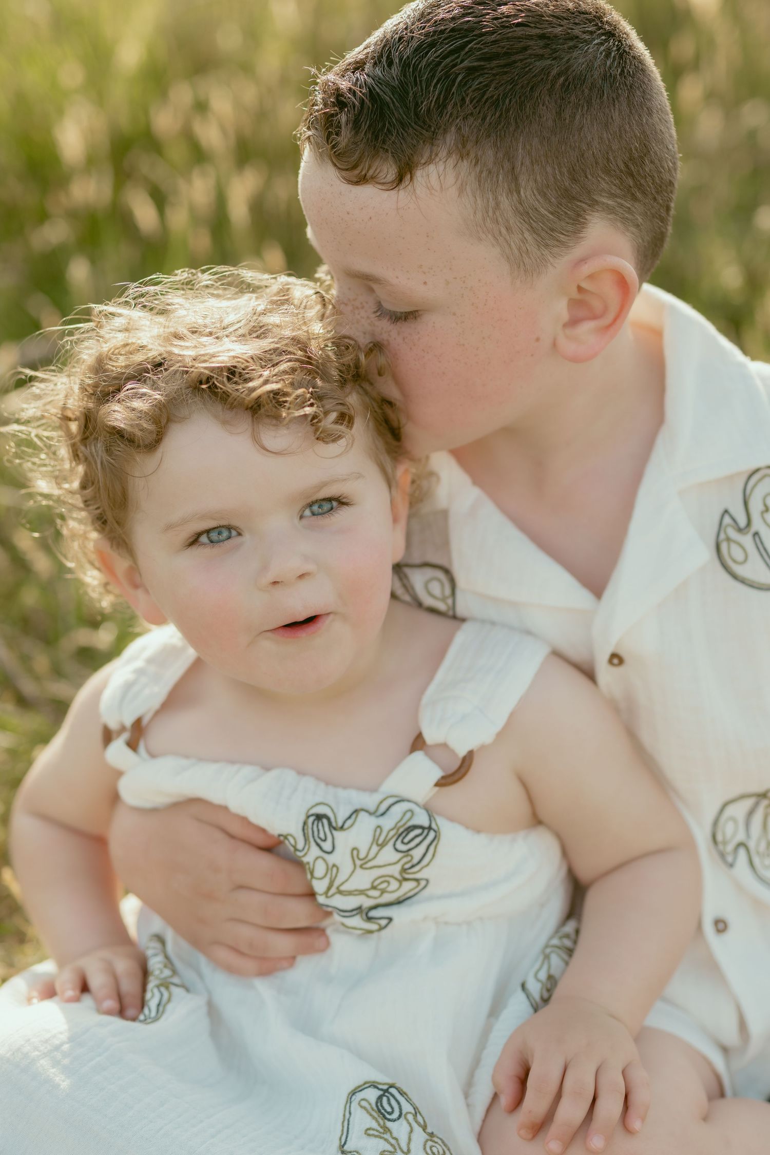 Two young children in white outfits playing together in sunlit field.