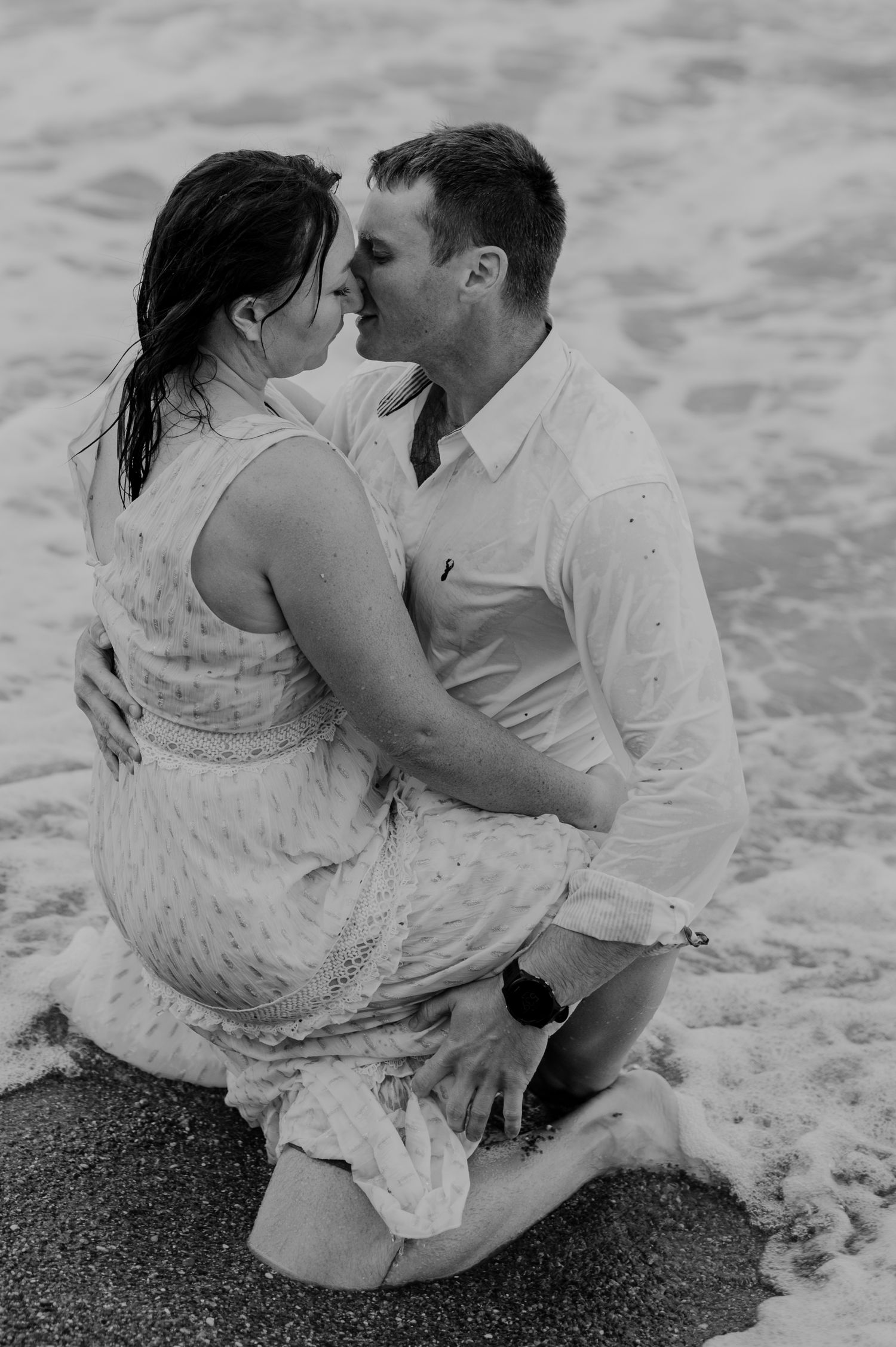 Black and white photo of a couple embracing in ocean waves on the beach.