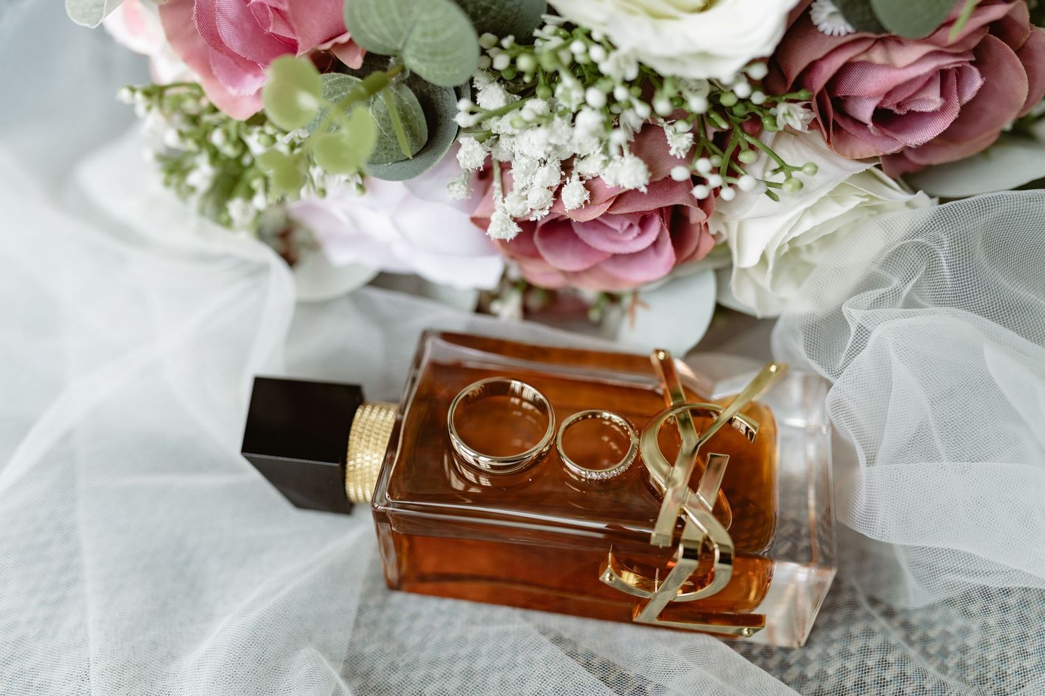Wedding rings rest elegantly on a perfume bottle surrounded by pink roses and delicate white tulle fabric.