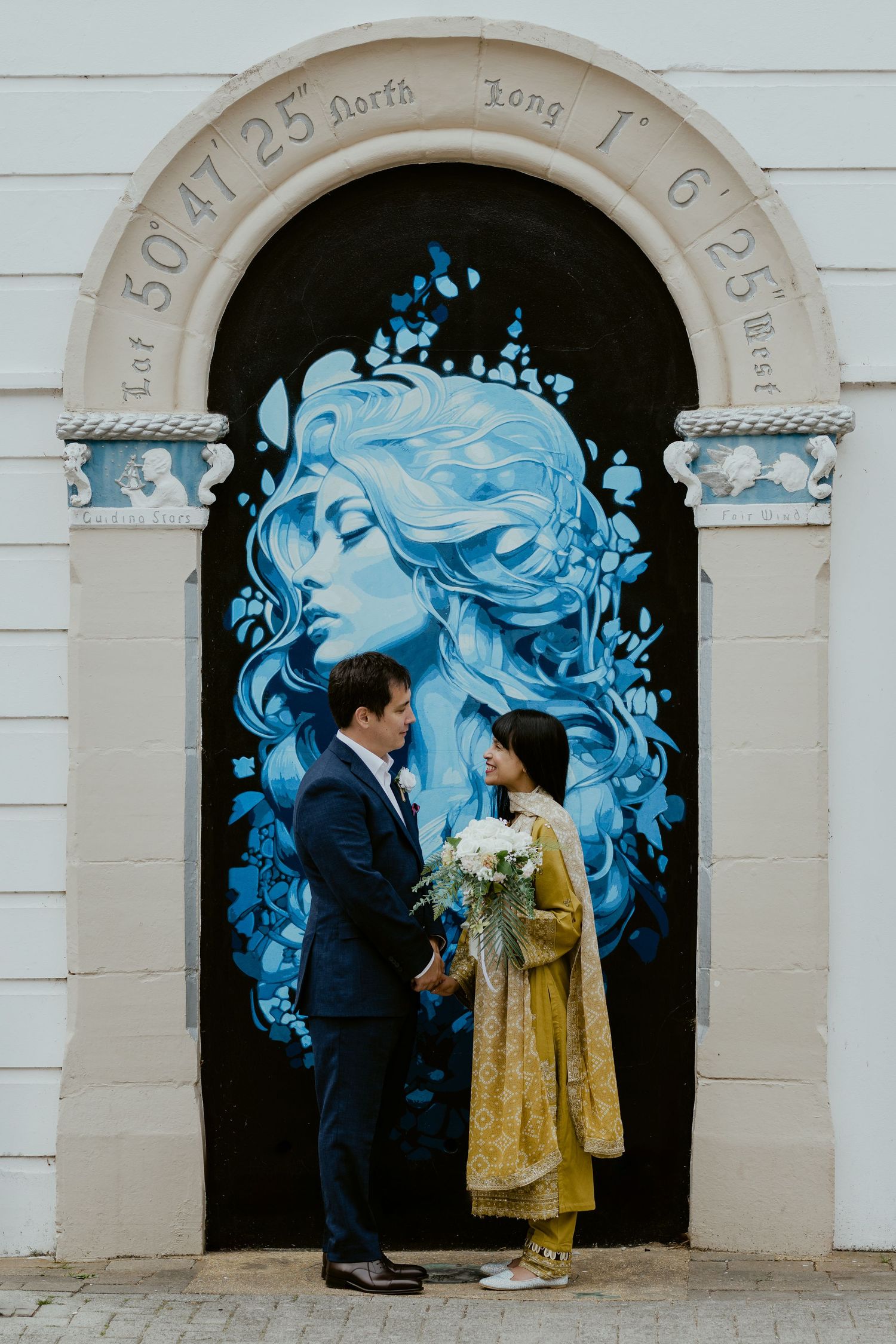 Couple standing against white wall with striking blue mural artwork in arched doorways.