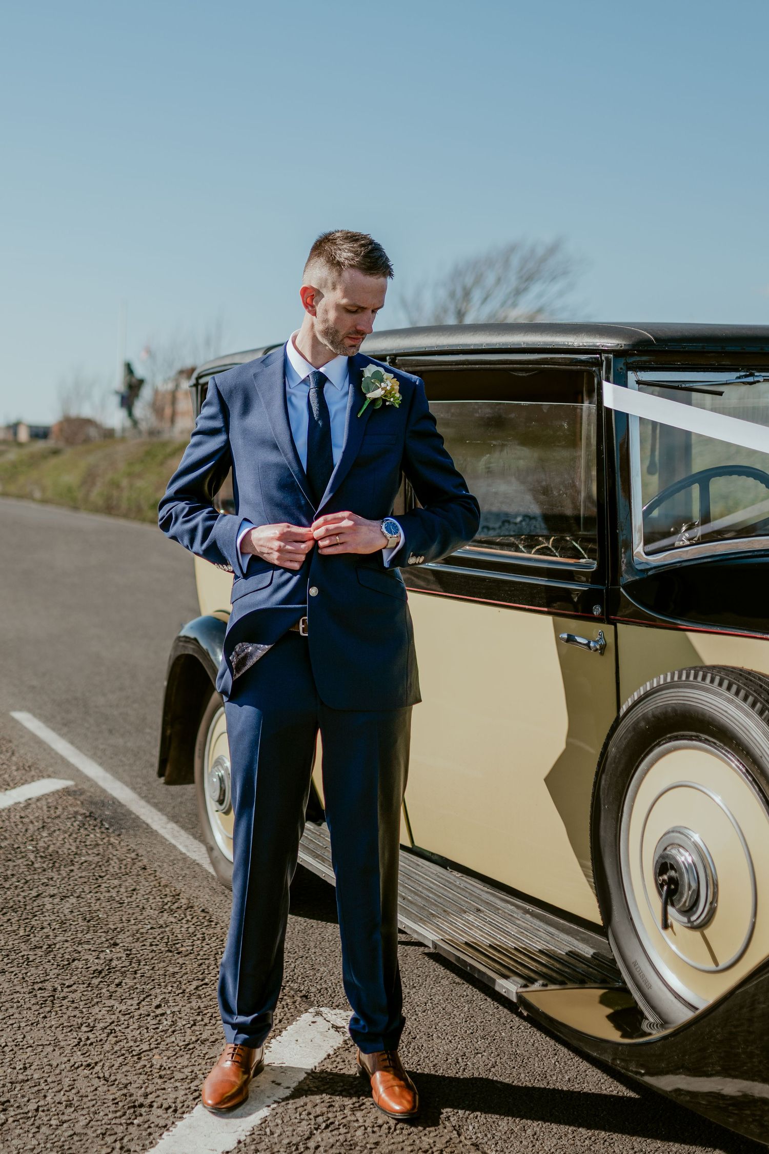 A person in a navy suit sits by a vintage cream-colored car.