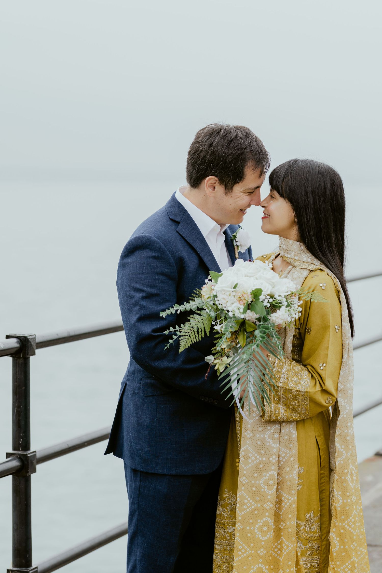 Couple embracing by waterfront railing with elegant floral bouquet.