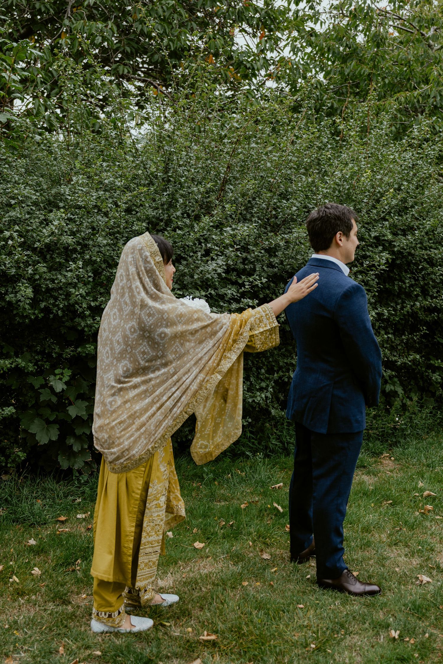 First look moment in a garden with bride in gold veil touching groom's shoulder.