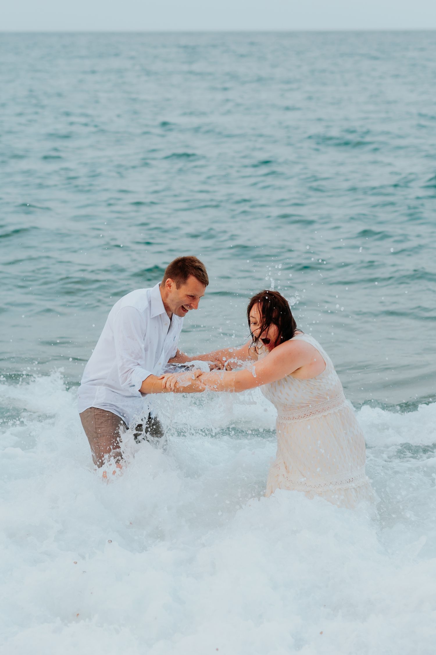 Couple playing and splashing in ocean waves wearing white wedding attire.