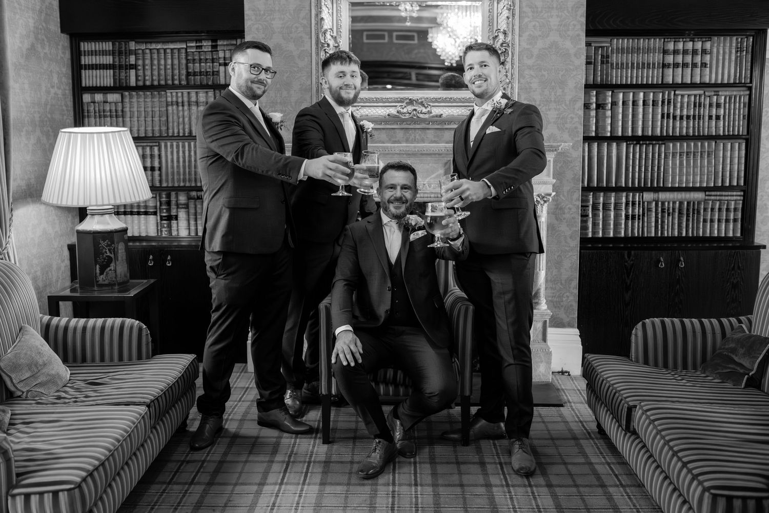 Groomsmen lounging in library with vintage furniture and bookshelves.
