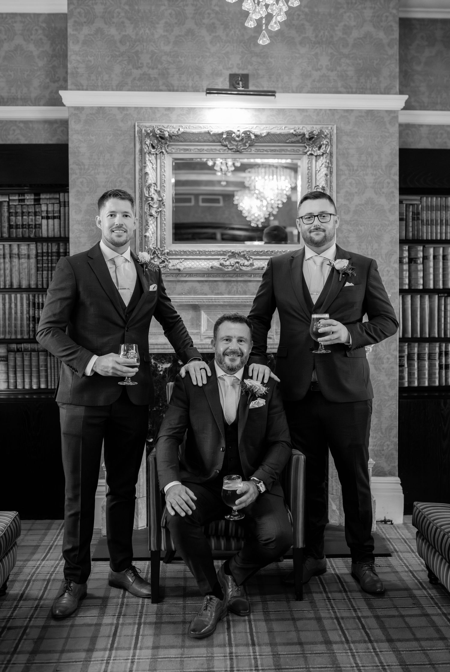 Groomsmen in black suits relaxing on vintage furniture in a library setting.