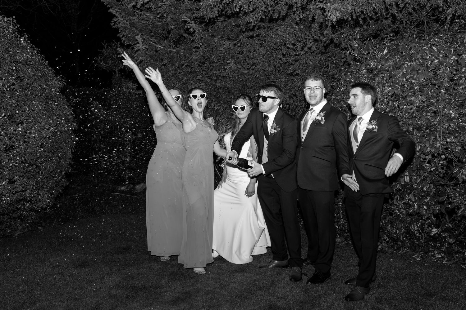 Wedding party members pose together in formal attire against a dark foliage backdrop.
