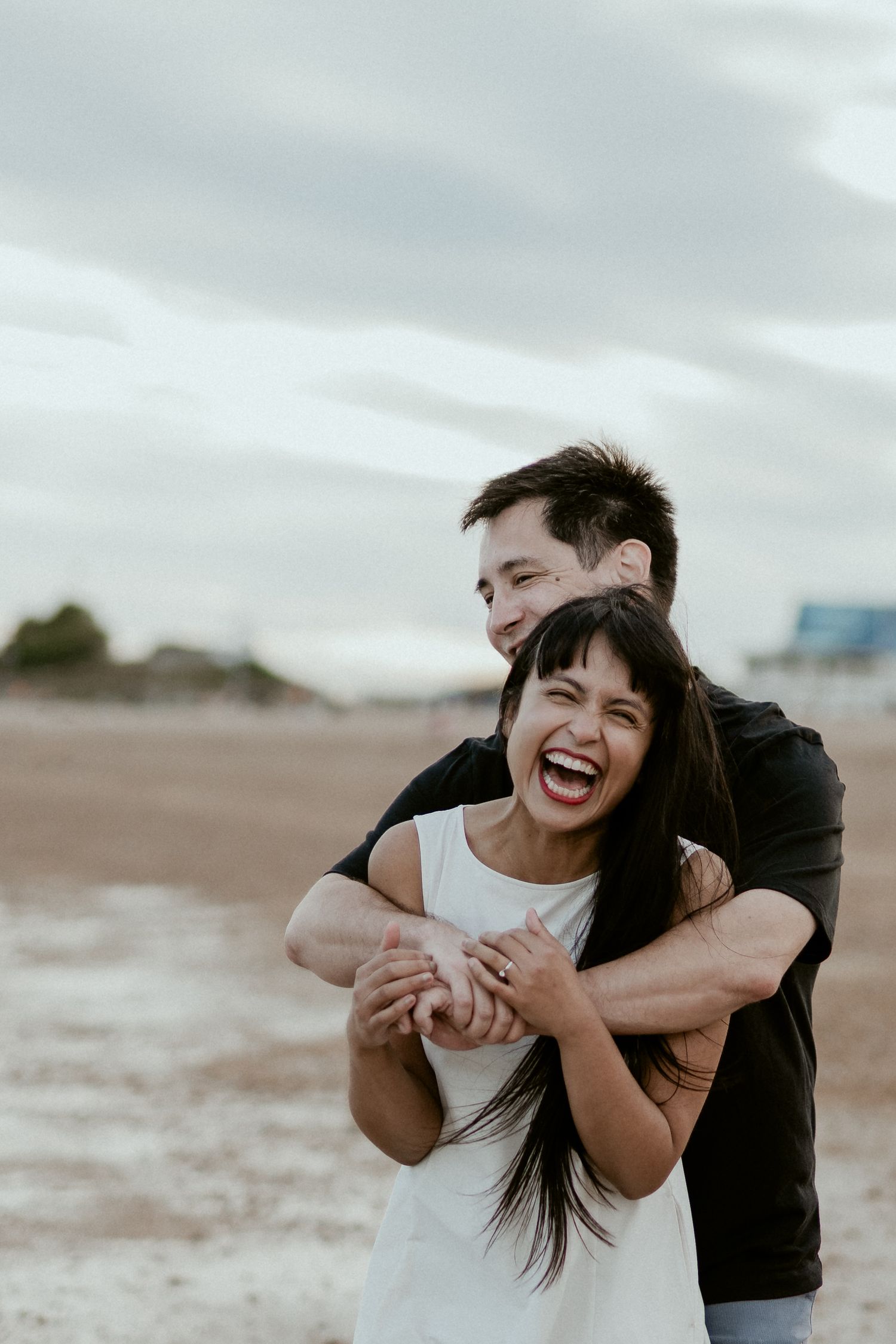 A couple shares a joyful embrace and laughter while standing on a beach under cloudy skies.