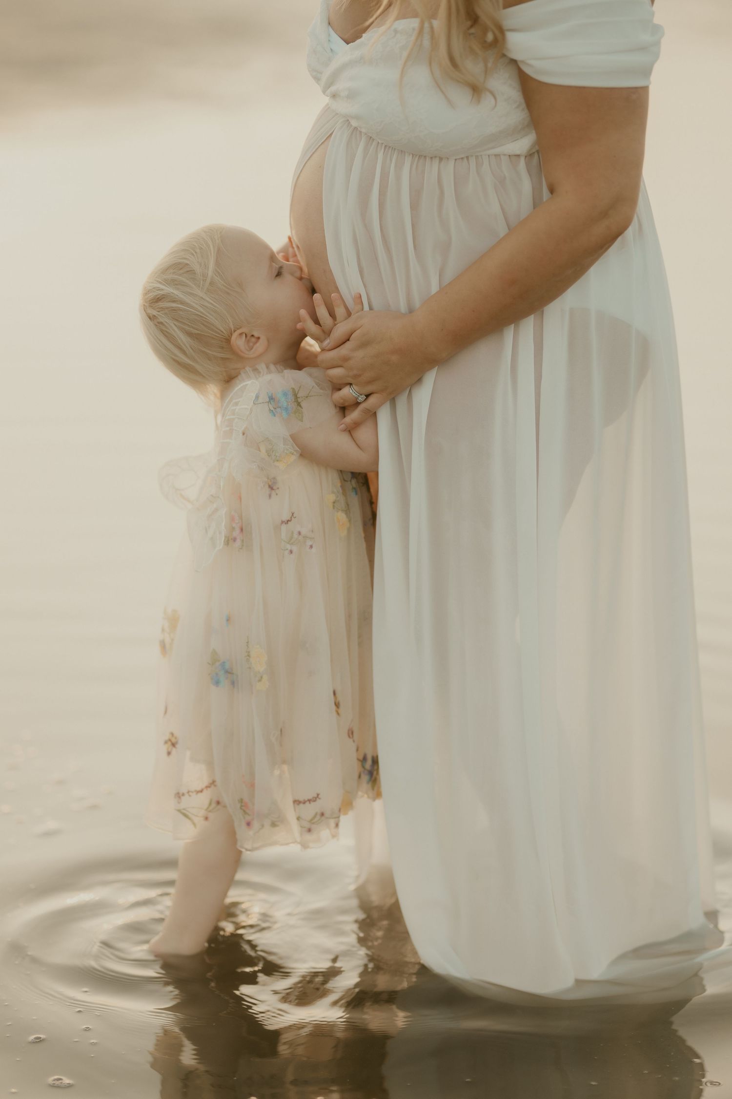 Parent and child in white dresses standing together in shallow beach water.