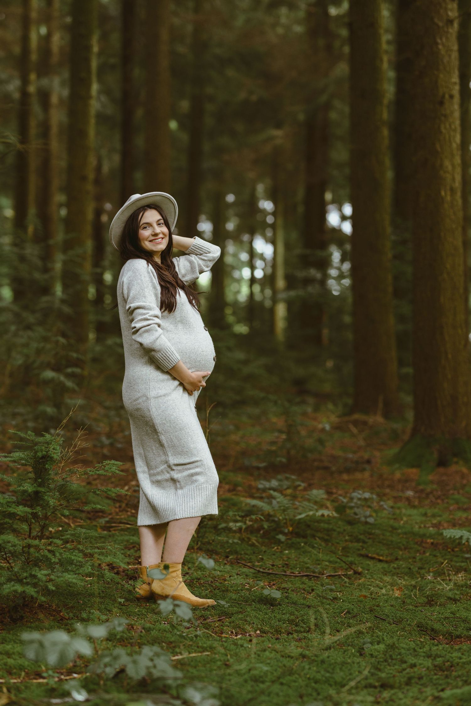 A person in a white sweater dress holds a small white dog in a serene forest setting.