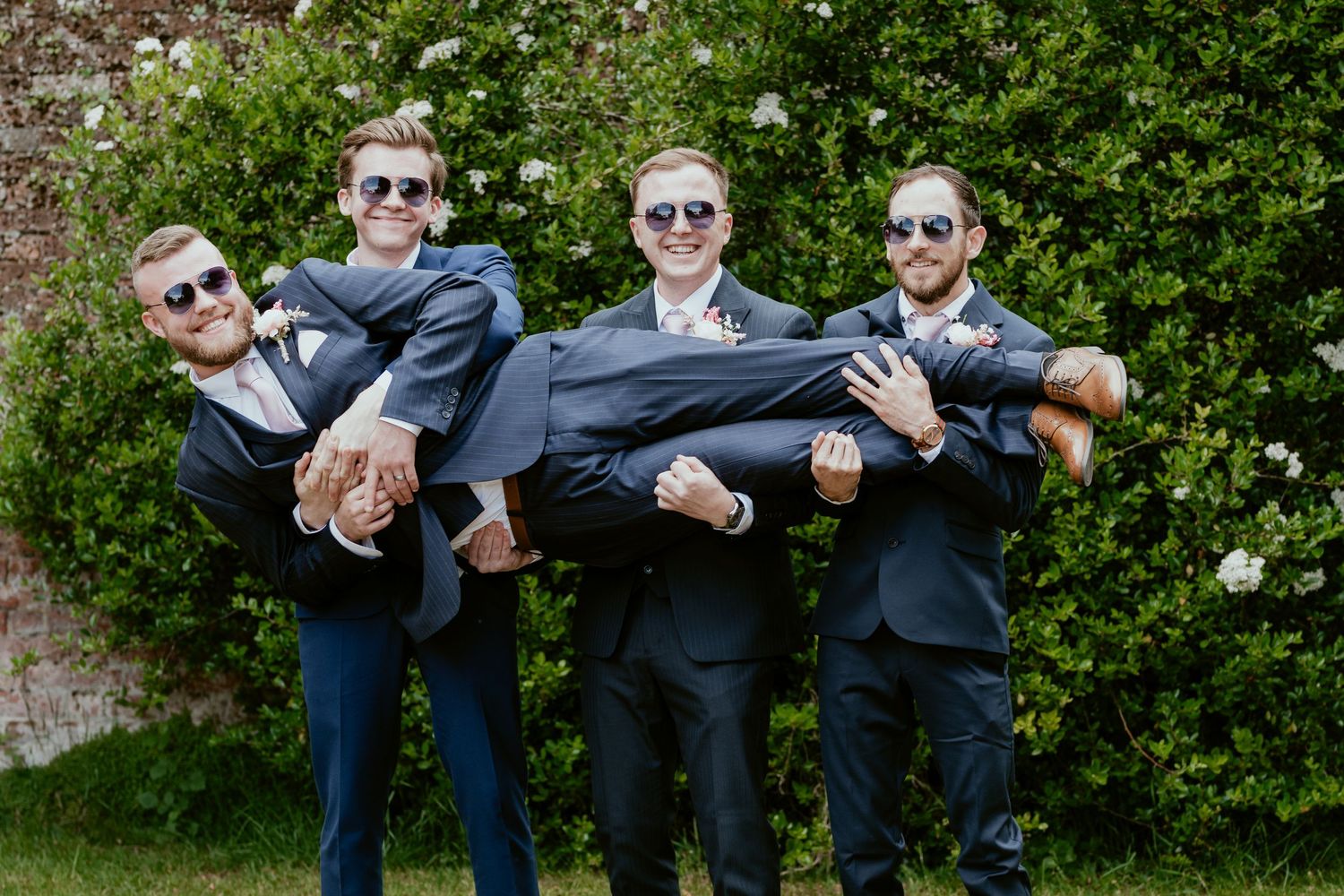 Groomsmen wearing sunglasses strike fun poses against green hedge backdrop.