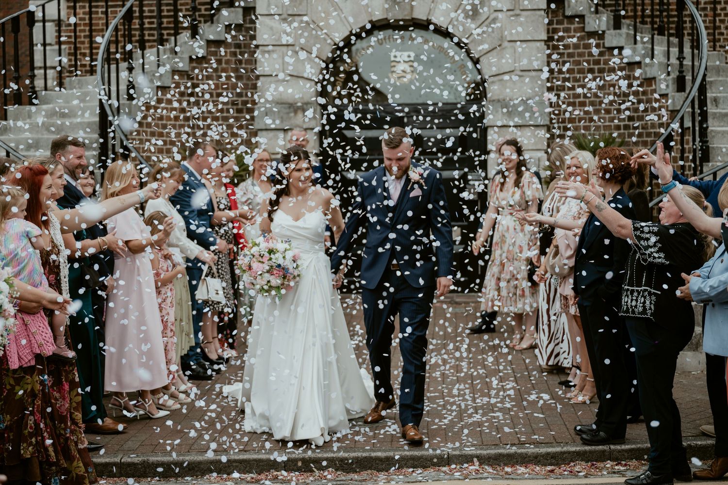 Wedding celebration with guests throwing confetti in front of a historic brick building.